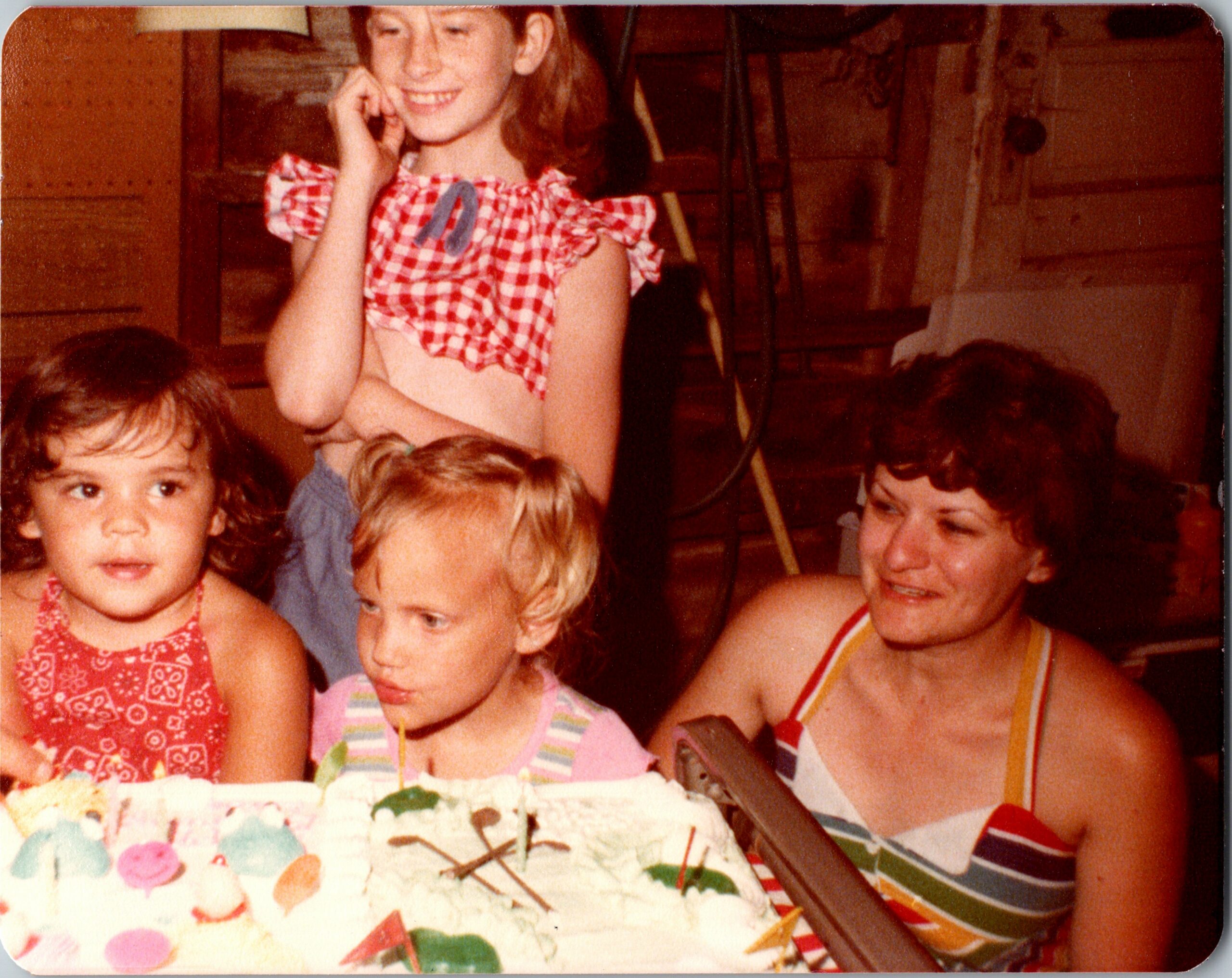 Children gathered around a decorated birthday cake with candles at a party, smiling and chatting indoors.