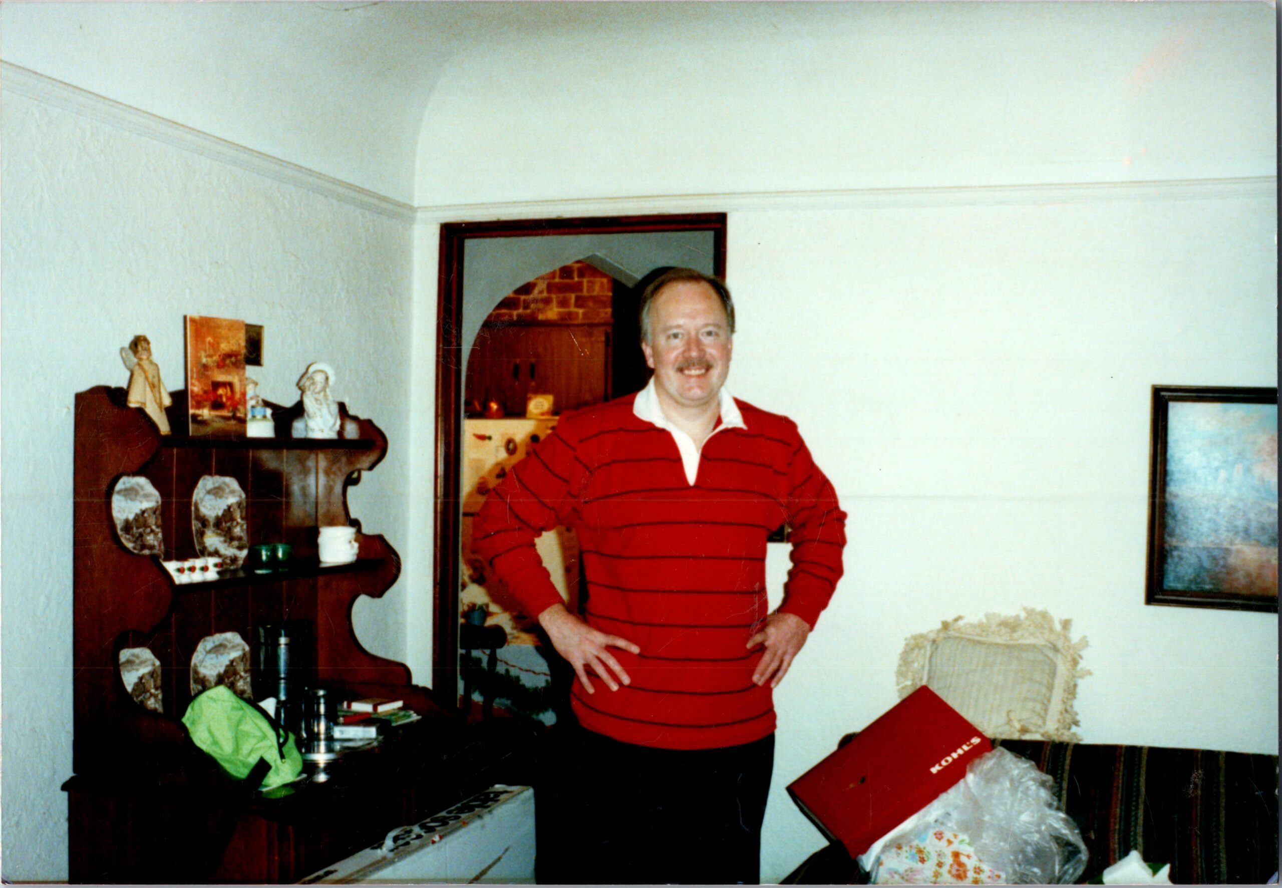 Smiling man in a red striped shirt stands in a cluttered living room with gifts and vintage furniture.