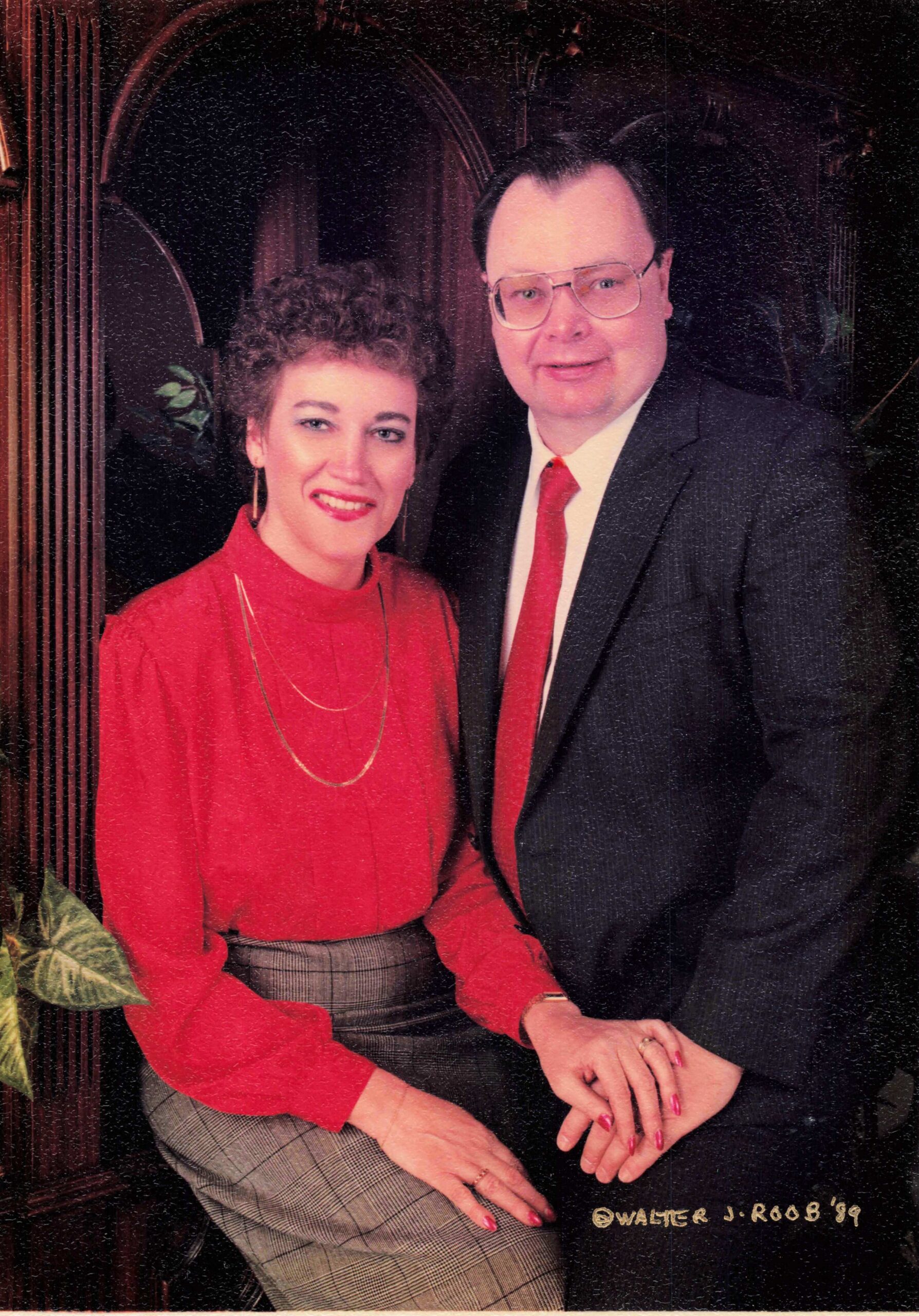 Smiling couple posing for a formal portrait: woman in a red blouse and plaid skirt beside a man in a dark suit with a red tie, seated together.