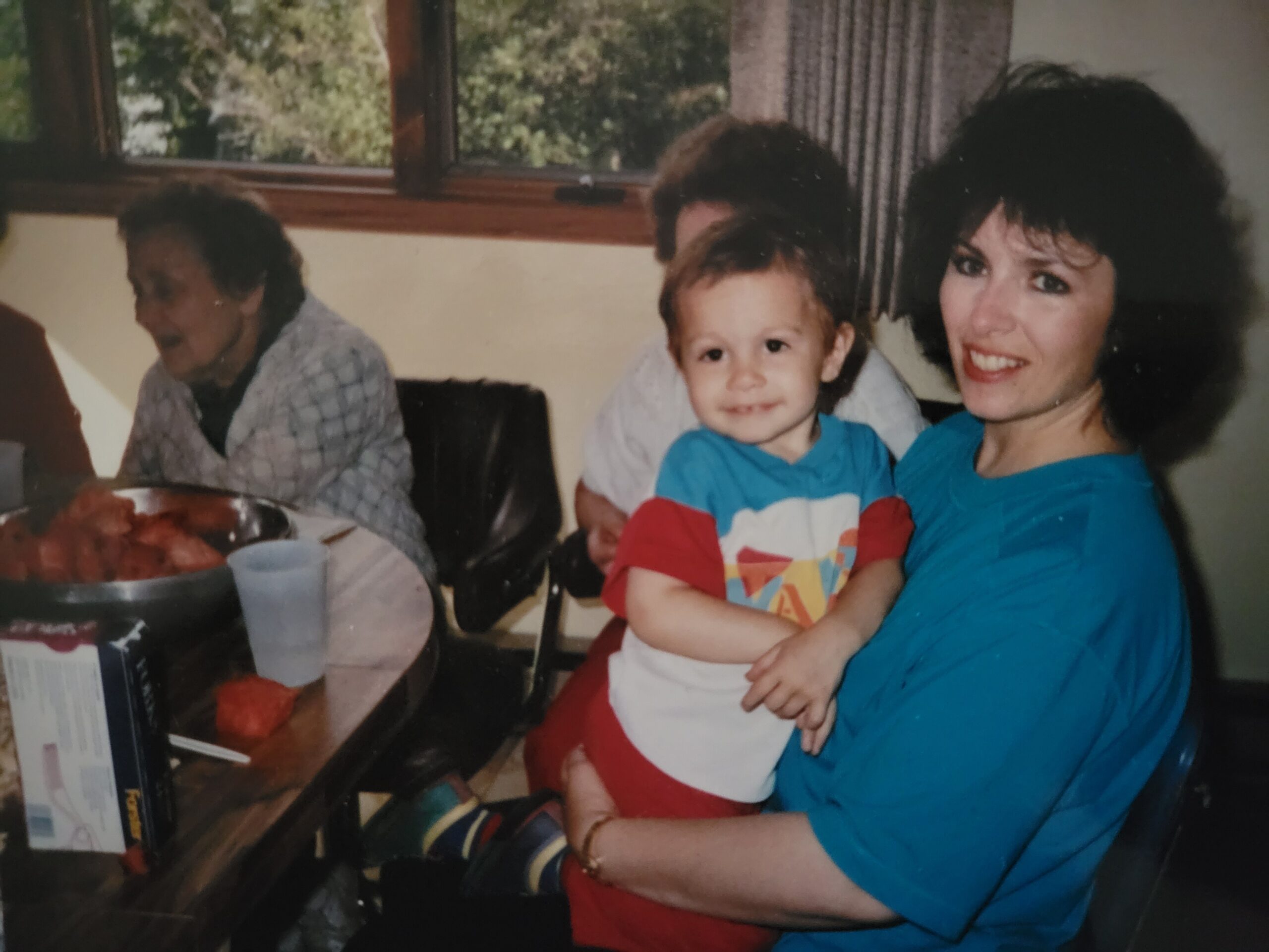 Smiling toddler in a blue shirt sits on a woman's lap at a dining table with food nearby in a home setting.