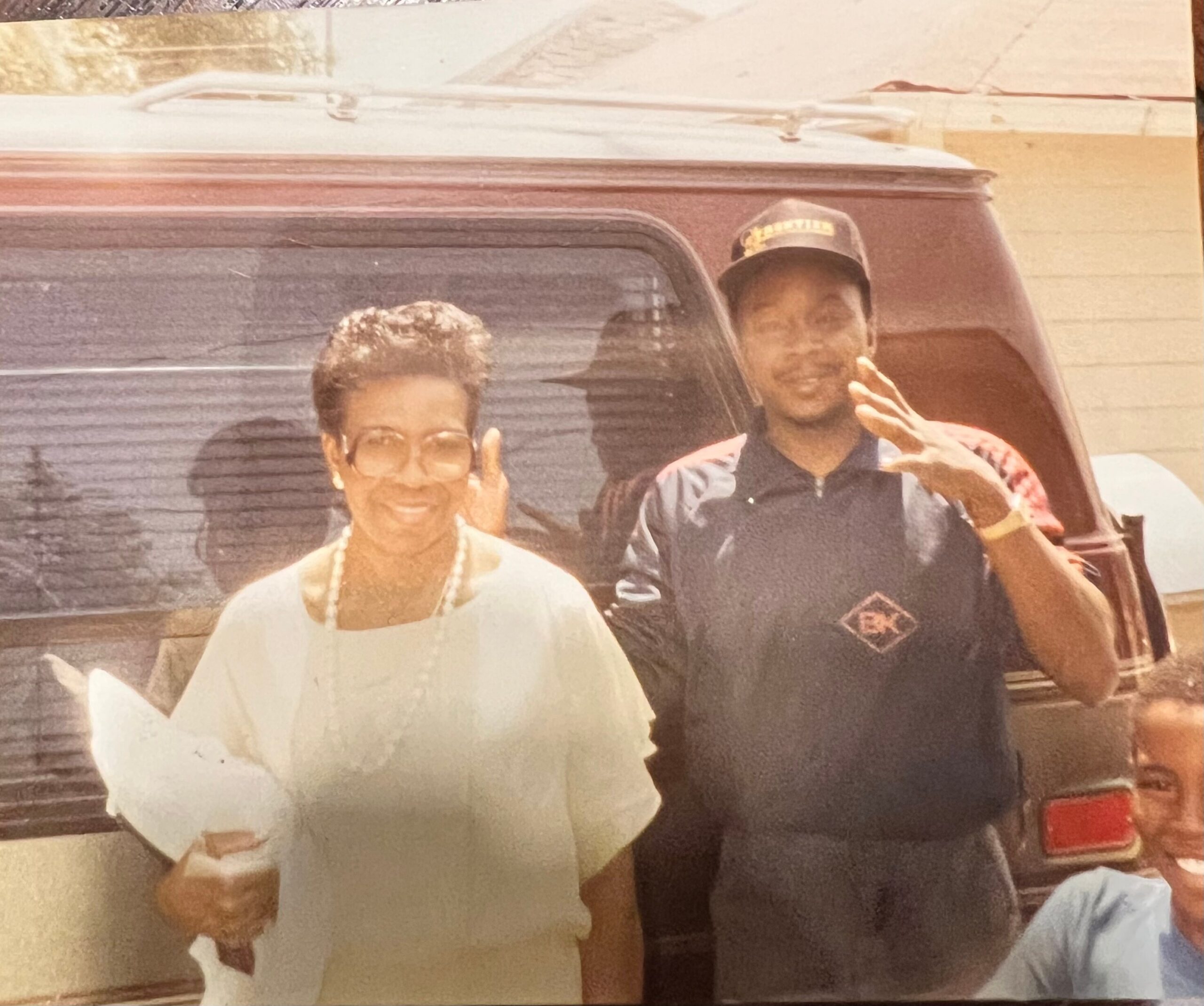 Two smiling people pose beside a maroon vehicle; man in a cap makes a peace sign while woman in light blouse stands next to him.