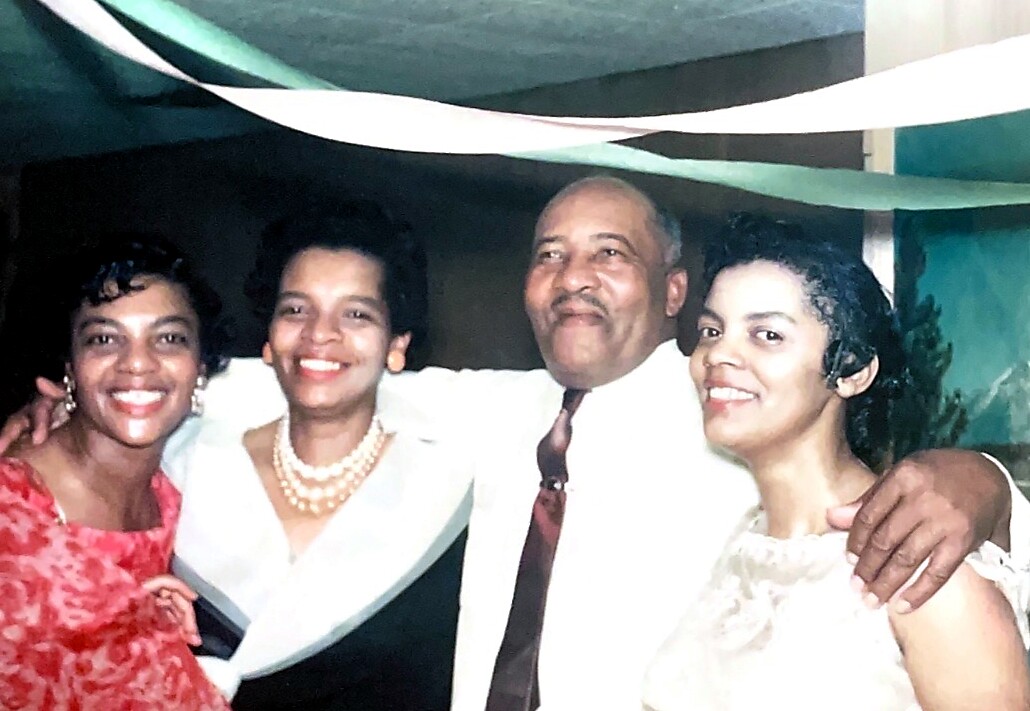 Four adults posing together at a celebration, smiling with streamers overhead in a festive setting.