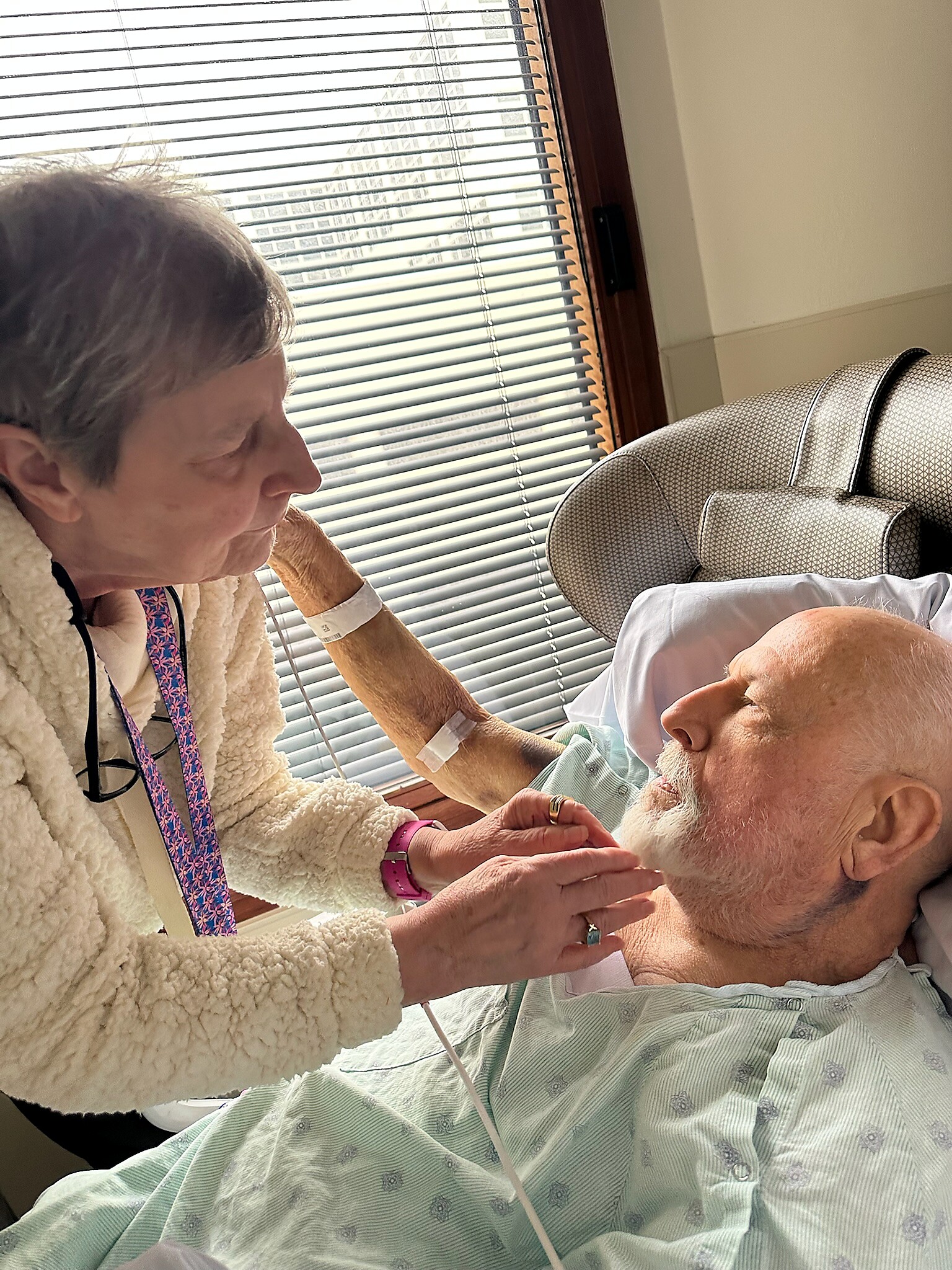 Caregiver helps an elderly man in a hospital bed, holding his hand near the headboard by a window with blinds.