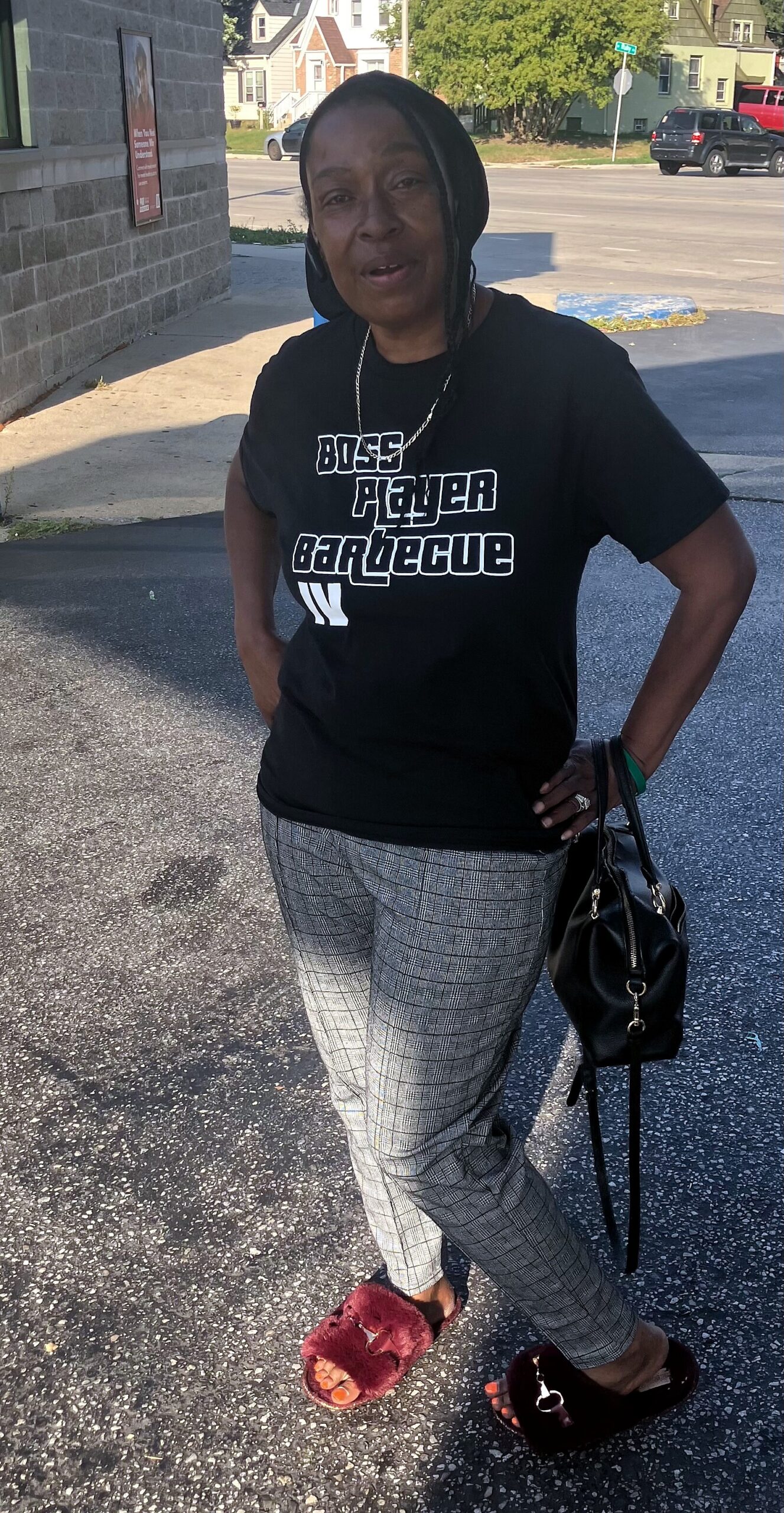 Woman standing outside wearing a black T-shirt that reads 'Boss Player Barbecue', gray plaid pants, red fuzzy slippers, and carrying a black handbag.