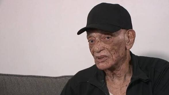 Elderly man in a black cap and dark shirt seated on a gray sofa, looking to the side.