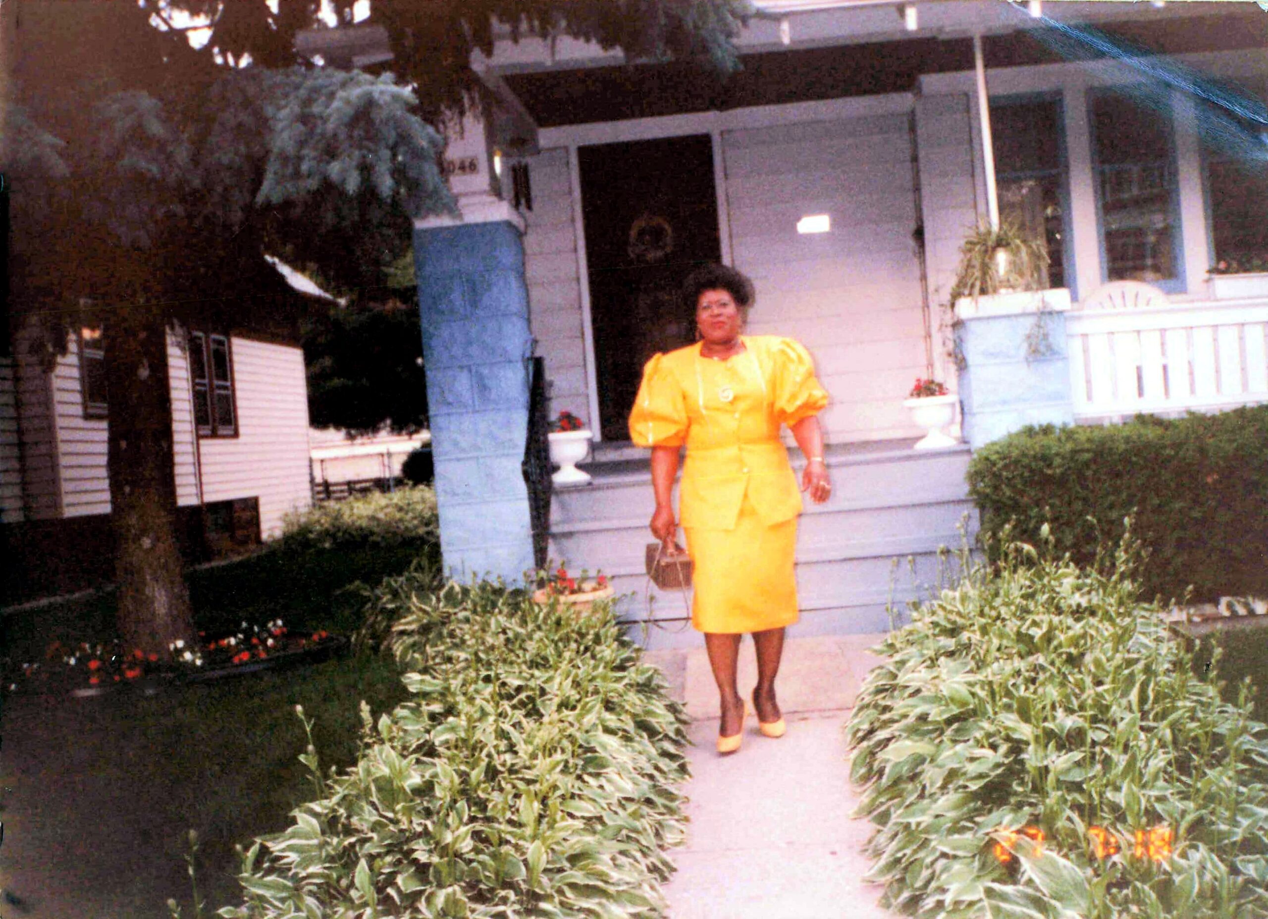 Woman in a bright yellow dress and matching shoes standing on the porch of a blue-and-white house with greenery around.