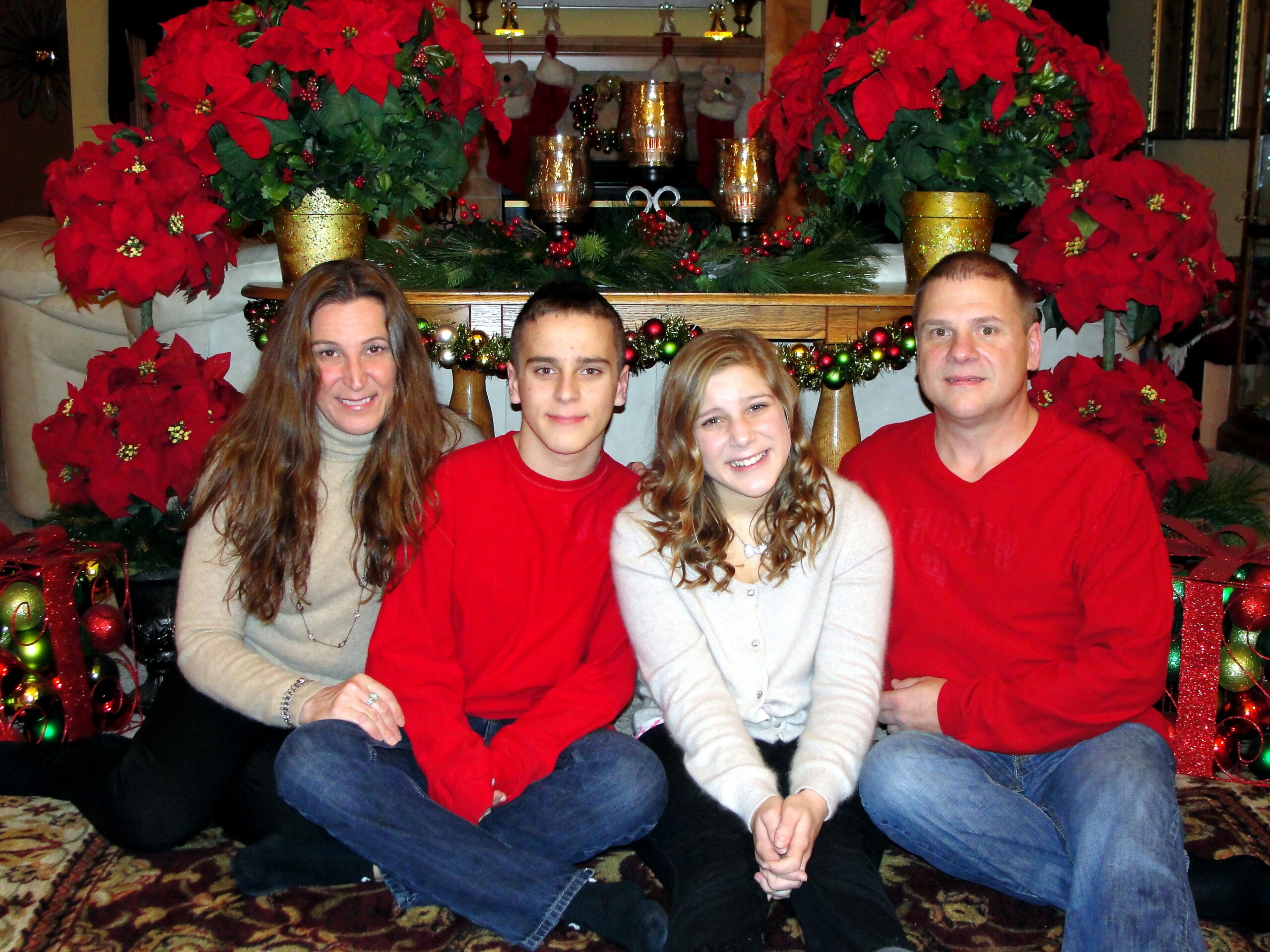 Family of four posing for a Christmas photo in front of a festive poinsettia display and garlanded altar.