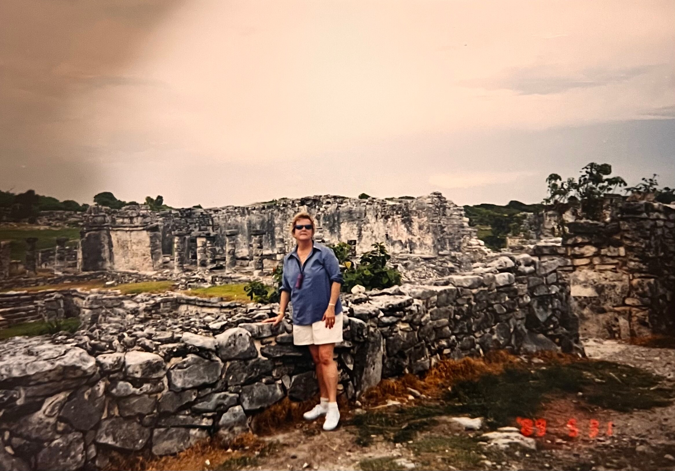 Woman wearing sunglasses in a blue shirt and white shorts standing among ancient stone ruins outdoors.