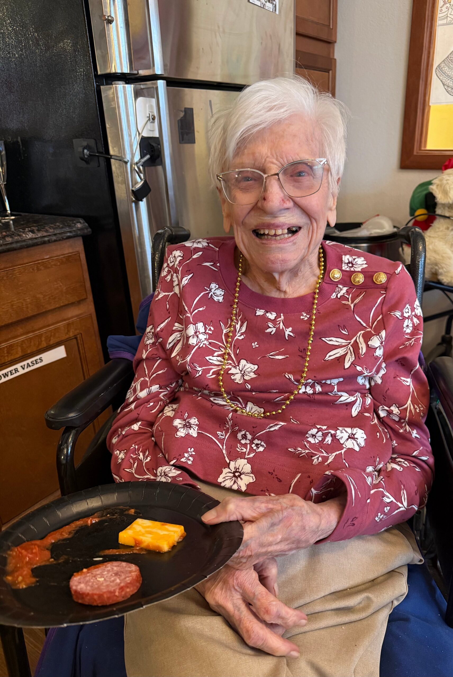Smiling elderly woman in a floral maroon sweater sits in a wheelchair at a table with a plate of food, including cheese slices and sausage, in a kitchen setting.