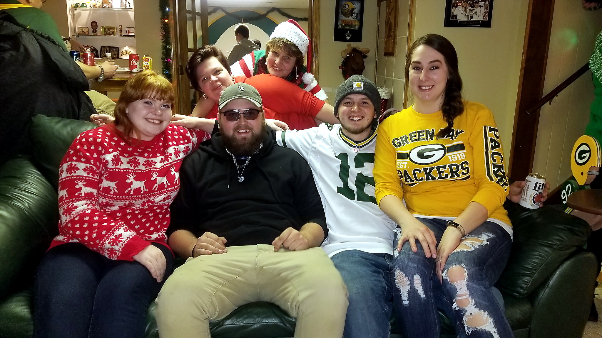 Six friends sit closely on a green leather couch in a festive room, wearing holiday sweaters and Green Bay Packers gear, smiling at the camera with drinks and holiday decor visible in the background.