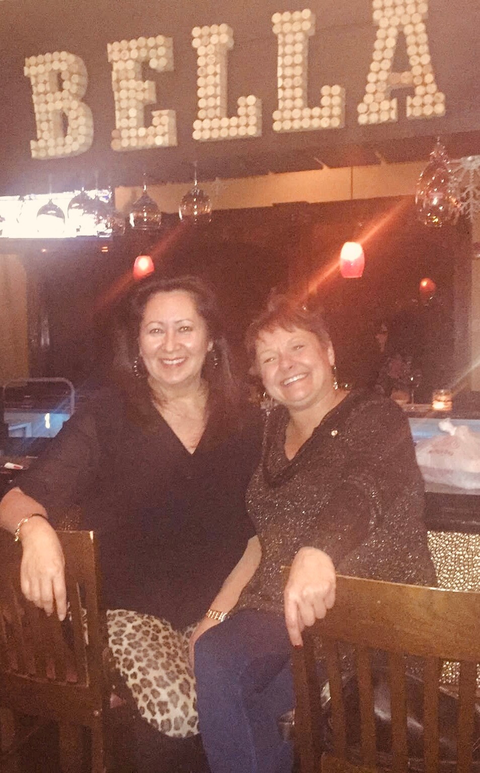 Two women sit together at a restaurant/bar, smiling for the camera under a large BELLA sign in the background.