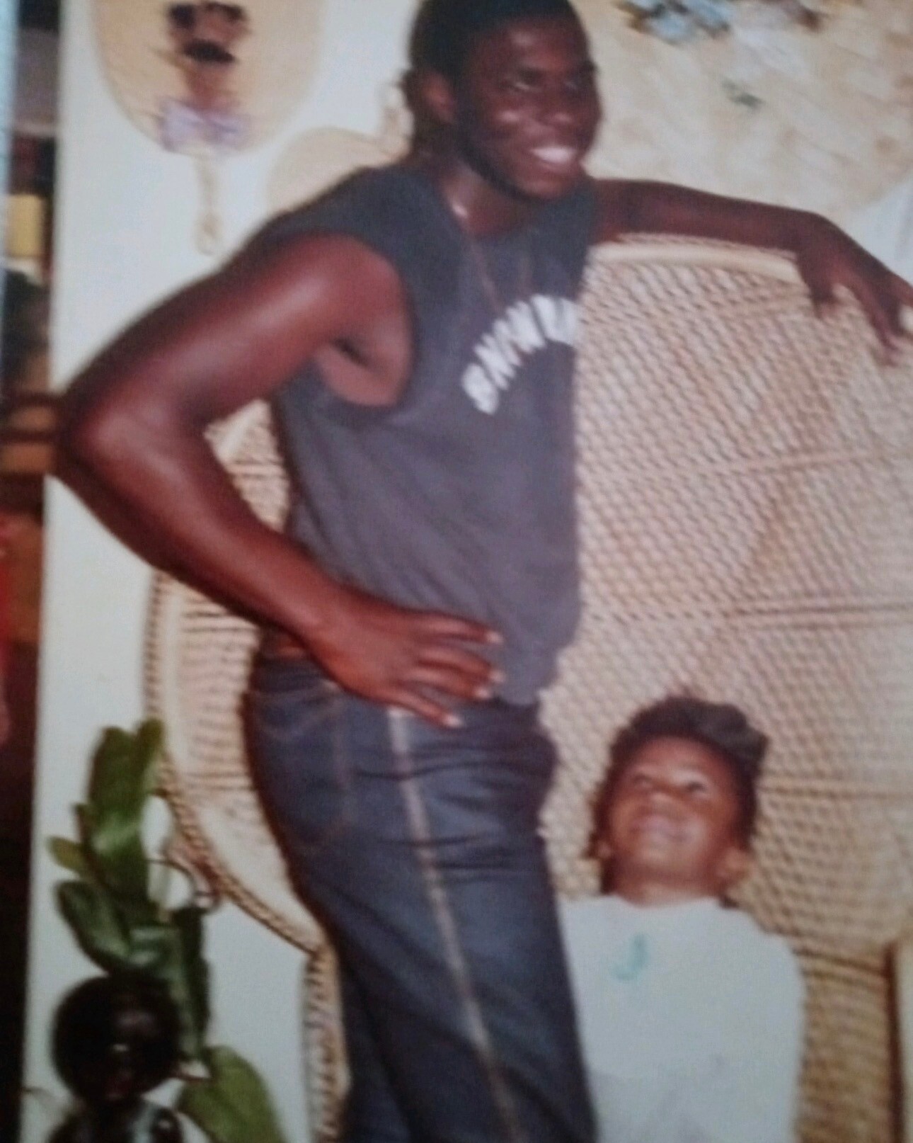 Smiling man in a dark sleeveless shirt leaning on a large woven chair indoors, with a child sitting on the floor nearby.