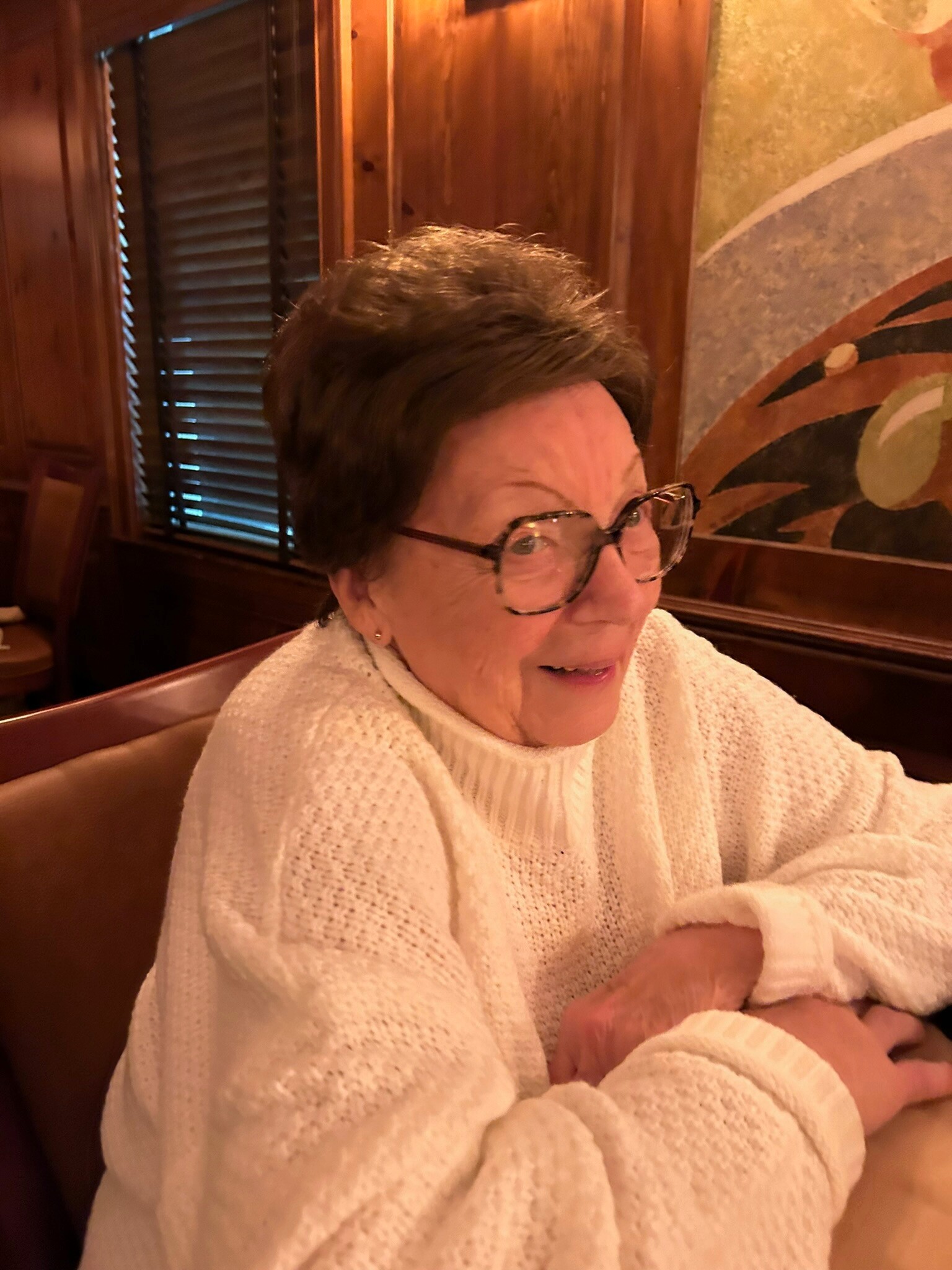 Elderly woman with short dark hair and glasses, wearing a white sweater, holding a newborn baby wrapped in a blanket.