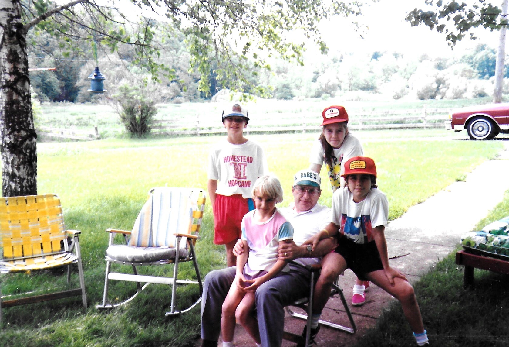 Group of children and adults posing outdoors on a grassy lawn with folding chairs, trees in the background and a red car nearby.