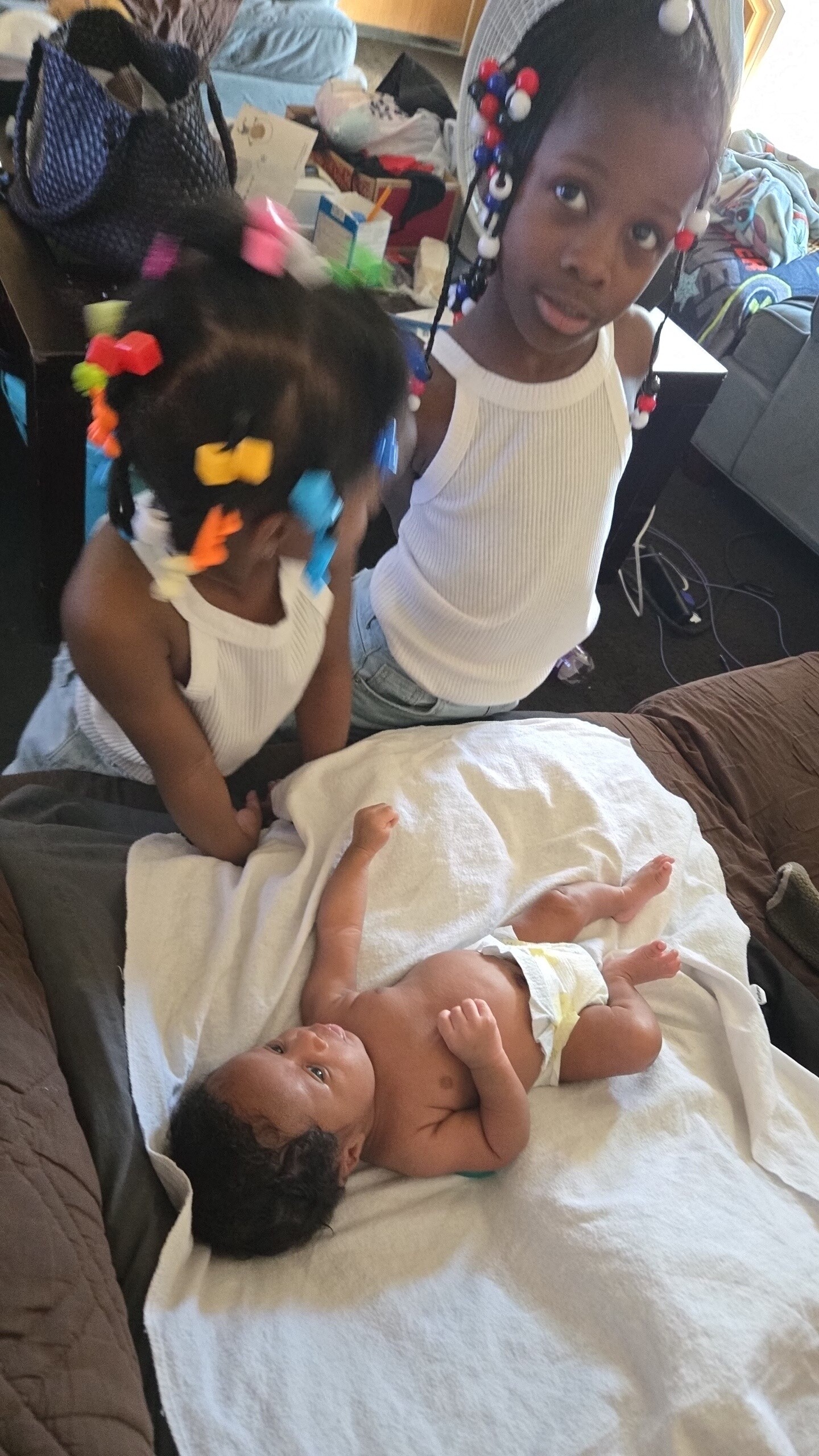 Two young girls with colorful beaded hairbands leaning over a baby lying on a white blanket in a bed or crib area in a cluttered room.