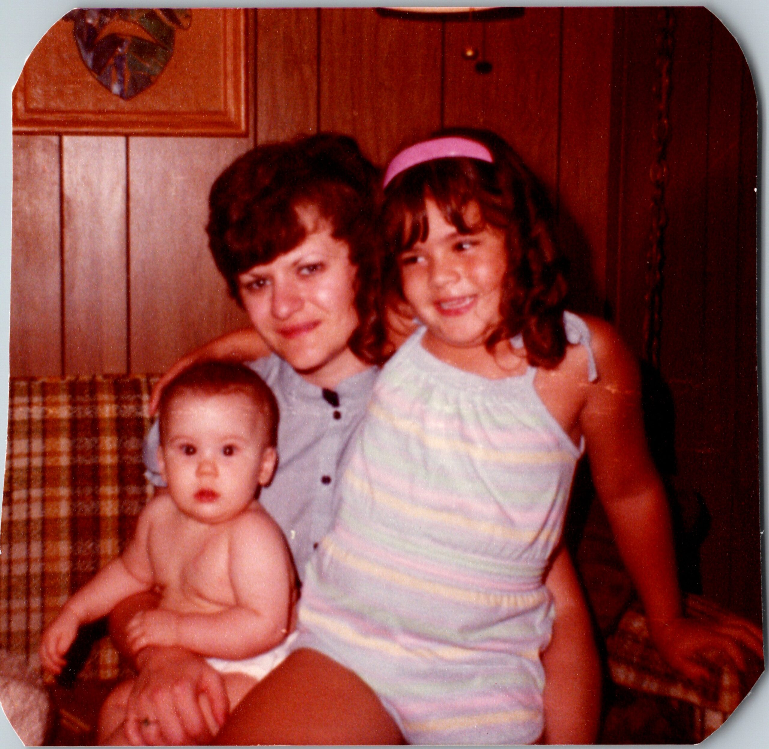 Three siblings posing closely for a family photo; a baby sits in front while two older children stand behind them indoors with wood paneling.