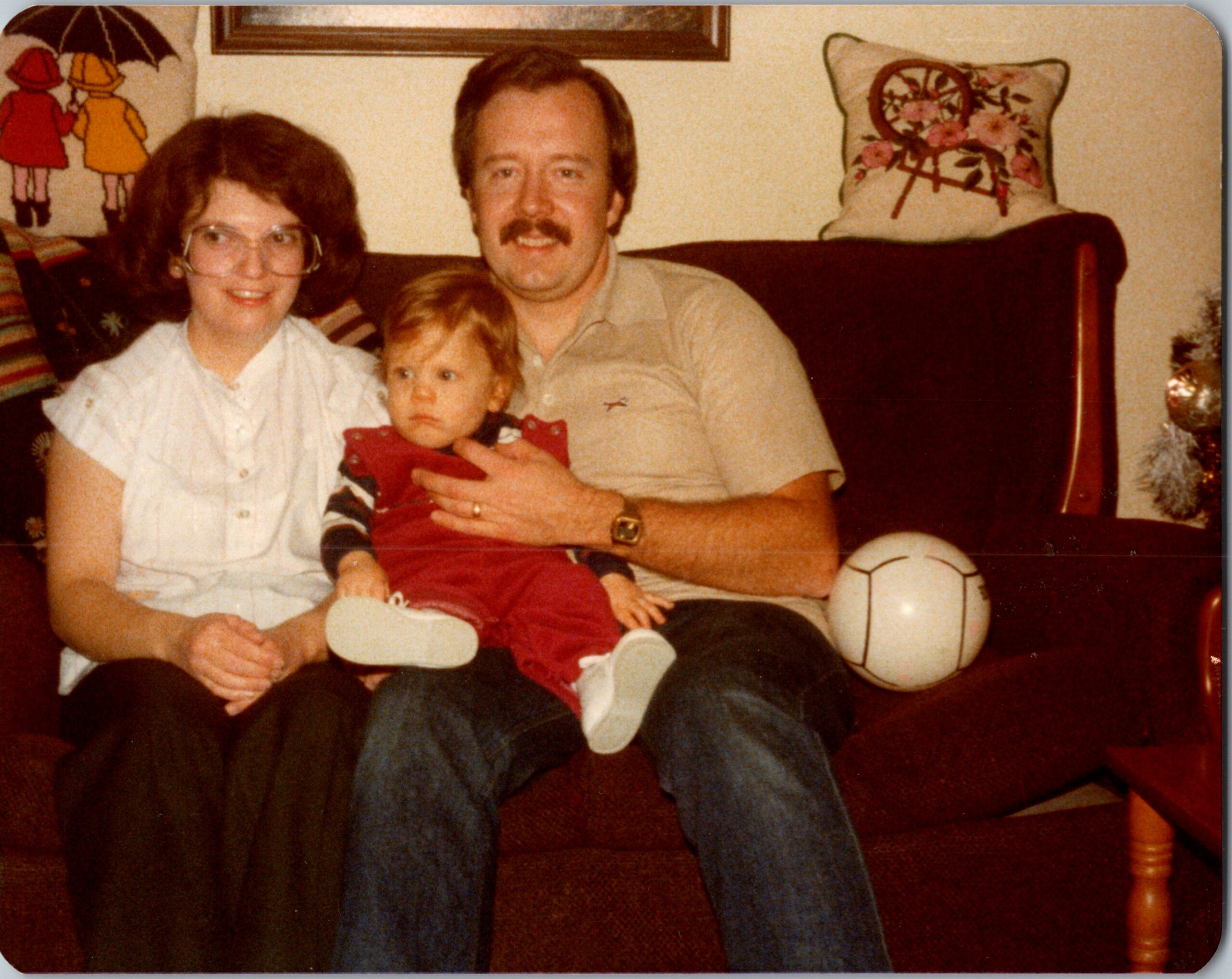 Family of three on a living room sofa: mother with glasses, father holding a toddler in red; a soccer ball nearby.