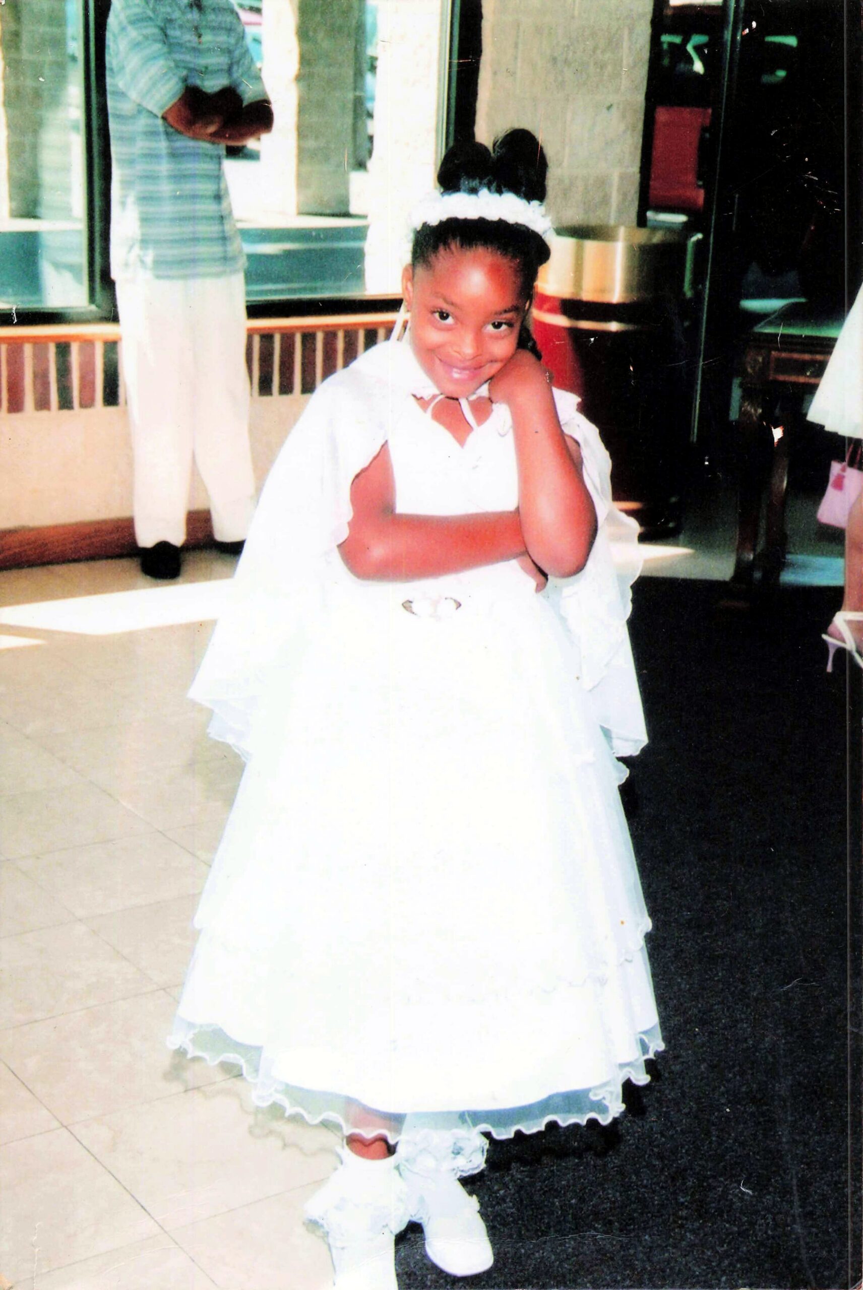 Young girl in a white, frilly dress with a fluffy headband, posing with her hand under her chin indoors at a party.