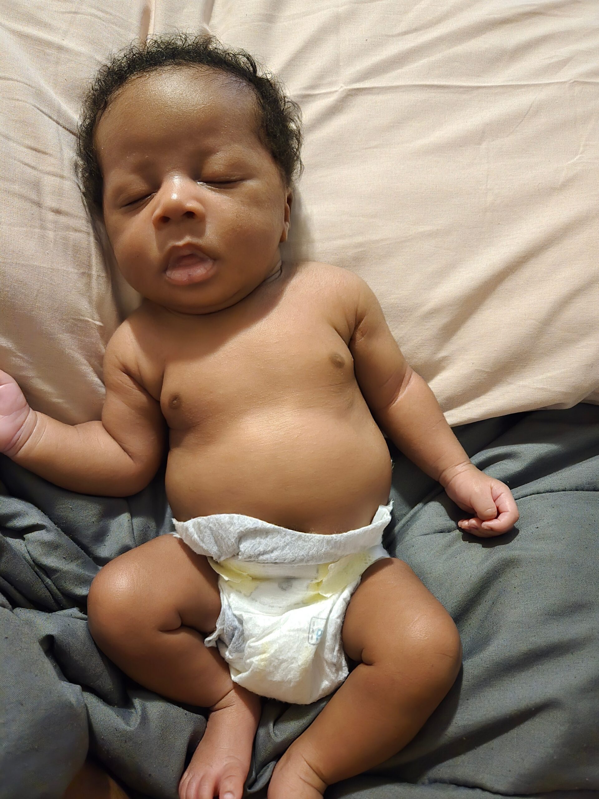 Infant sleeping on a beige sheet, eyes closed, wearing a diaper and bare torso visible from chest to hips.