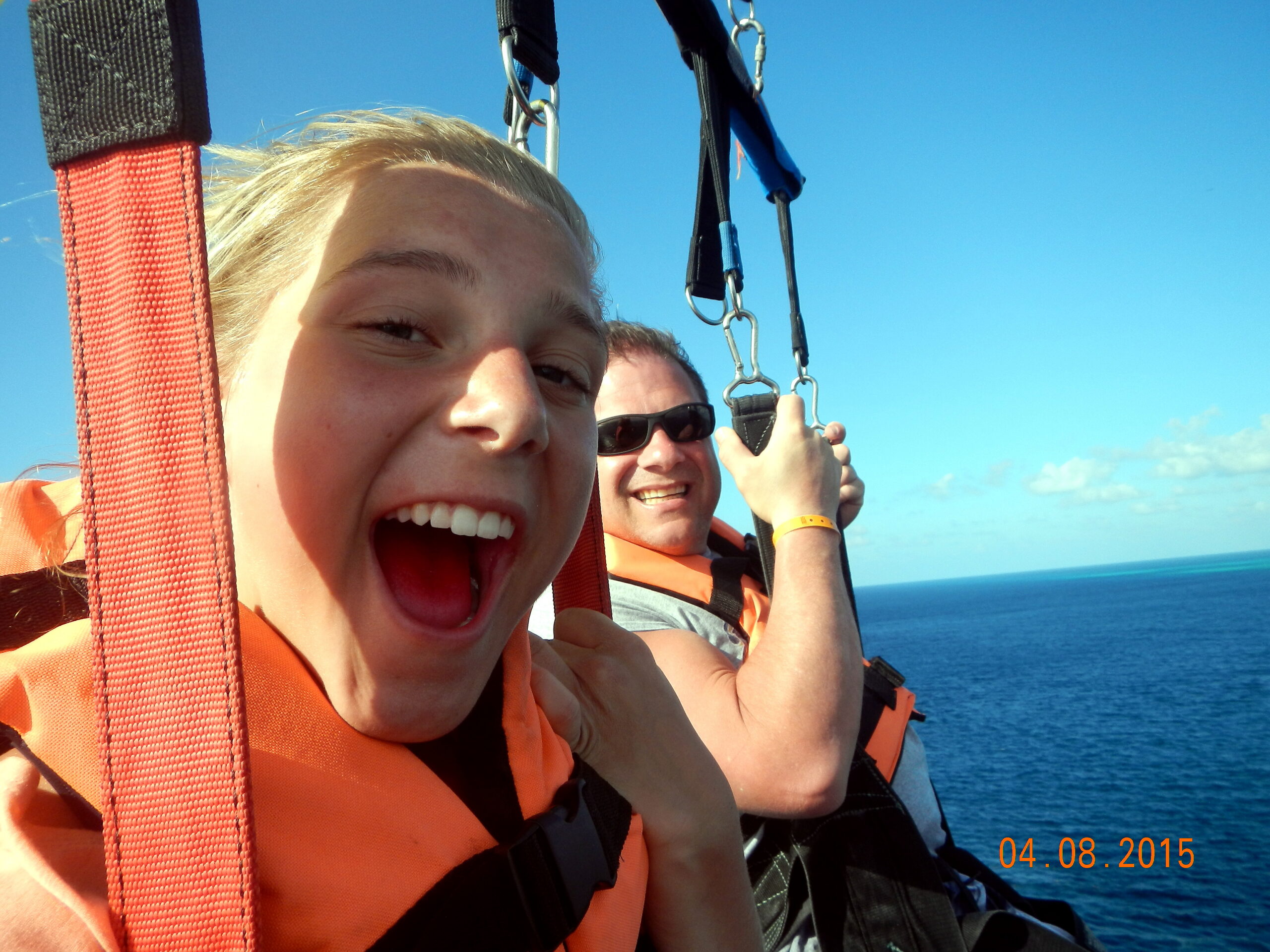 Two people parasailing over a blue ocean, wearing orange life jackets and smiling at the camera with harnesses visible.
