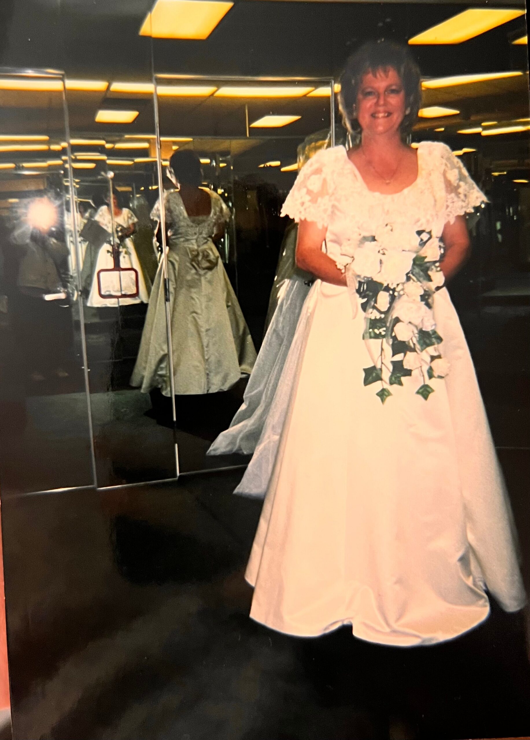 Bride in a white lace wedding gown smiling, holding a cascading bouquet of white flowers and ivy in front of mirrored walls.