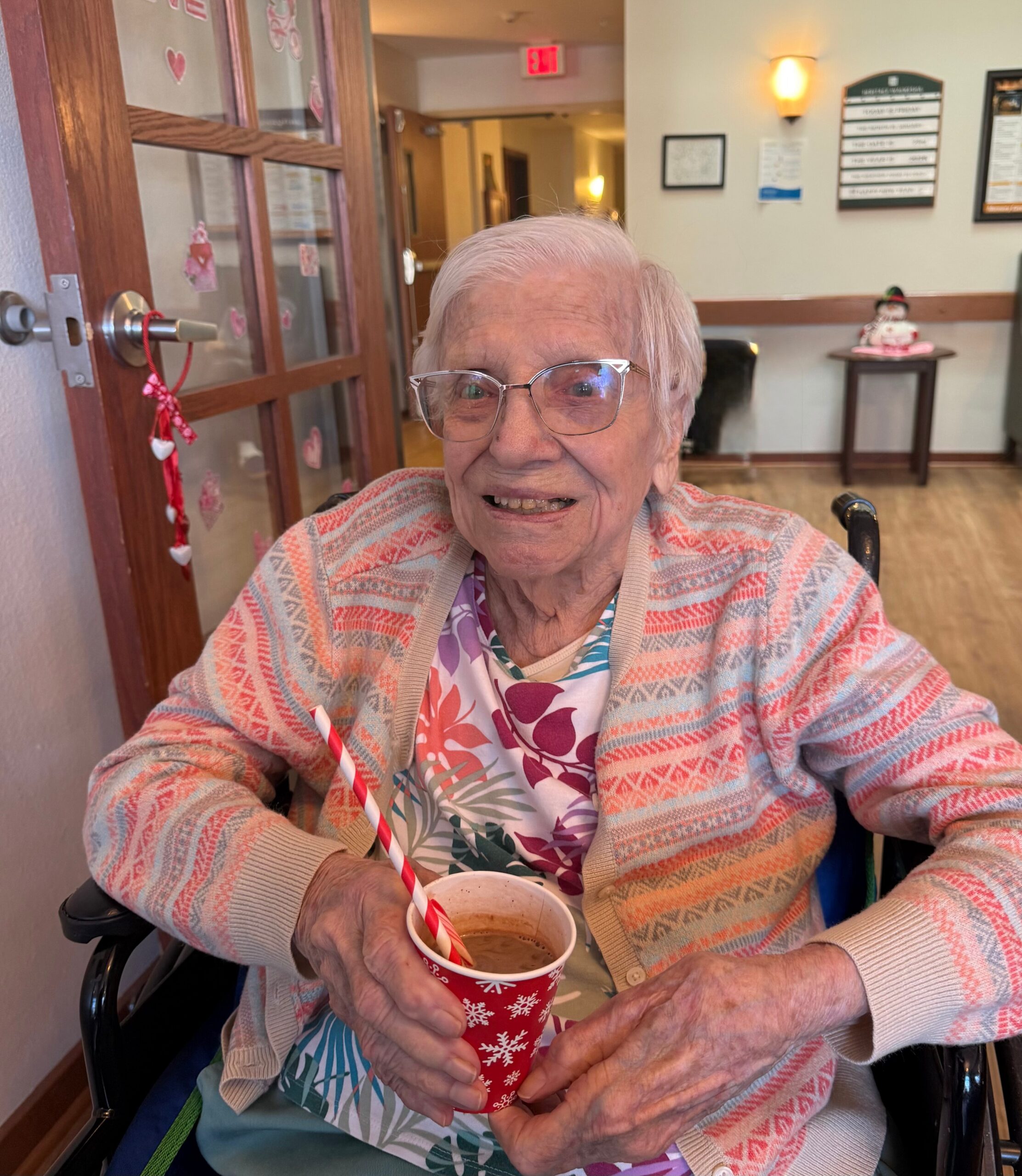 Smiling elderly woman in a wheelchair holding a red holiday cup with a striped straw, indoors with Valentine decorations on the glass door behind her.