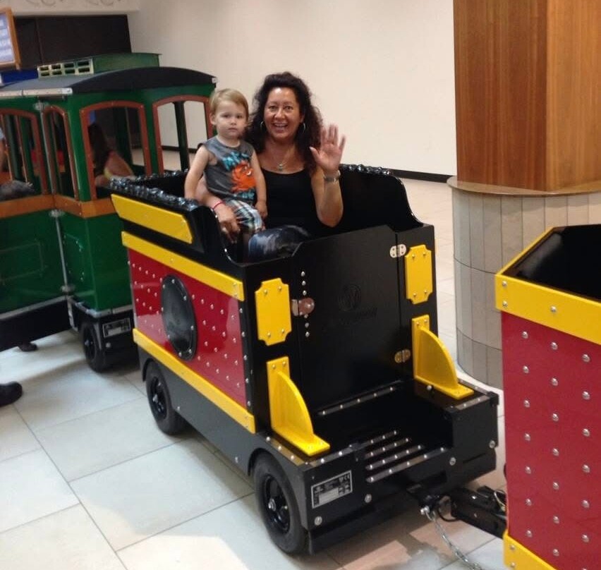 Smiling woman waves from a small colorful train car with a young child aboard in a mall setting.