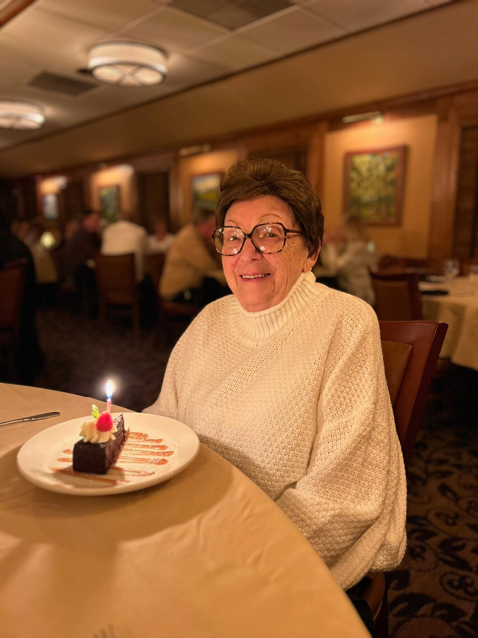 Older woman with short dark hair and glasses smiles at a restaurant table with a small chocolate cake and candle in front of her.