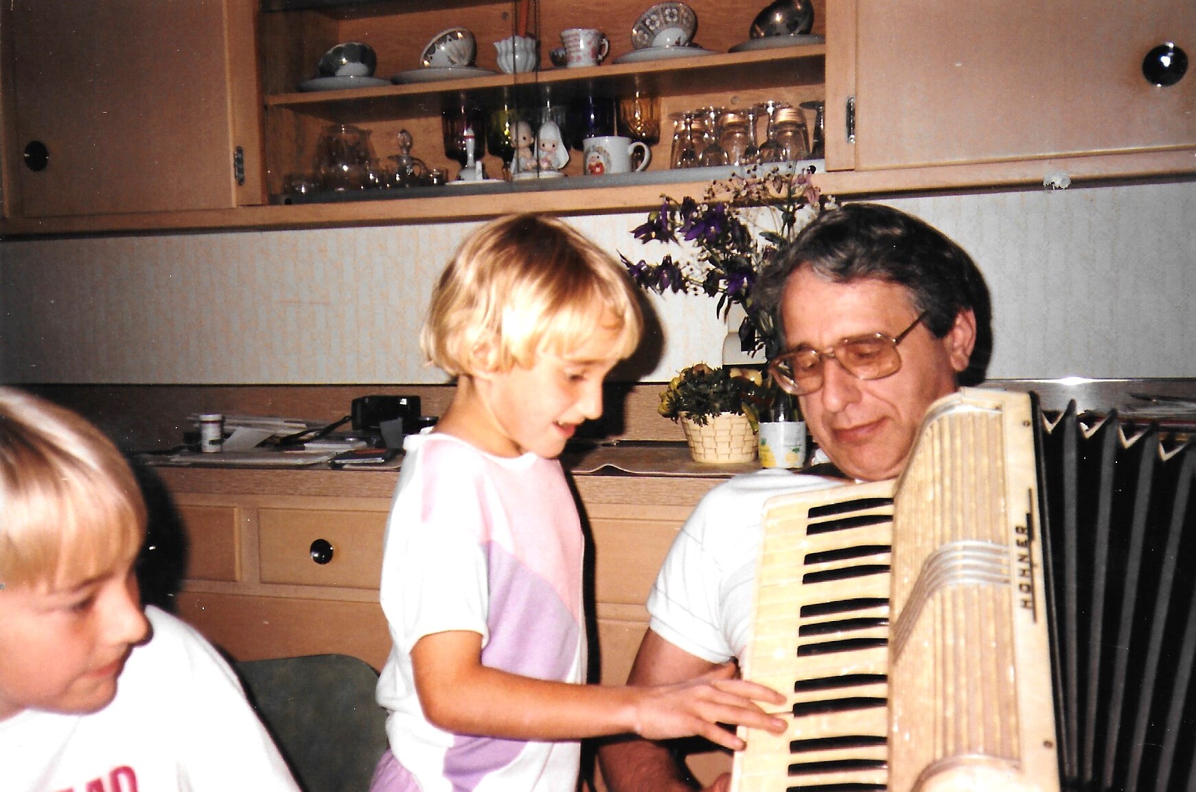Girl standing beside a man playing a large accordion in a home kitchen with wooden cabinets and dishes visible in the background, smiling.