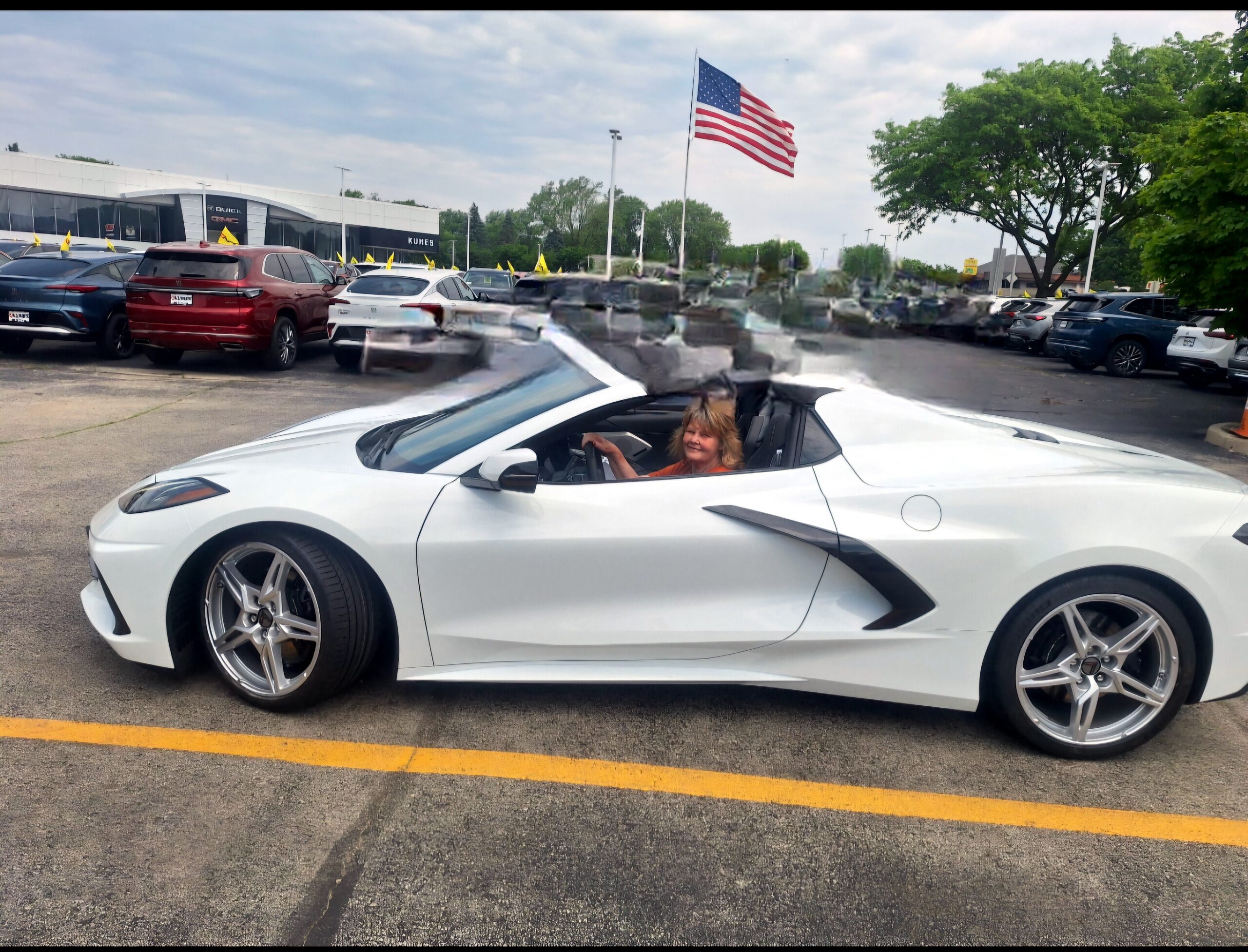 White convertible with a driver in the foreground, parked in a dealership lot with other cars and a waving American flag in the background