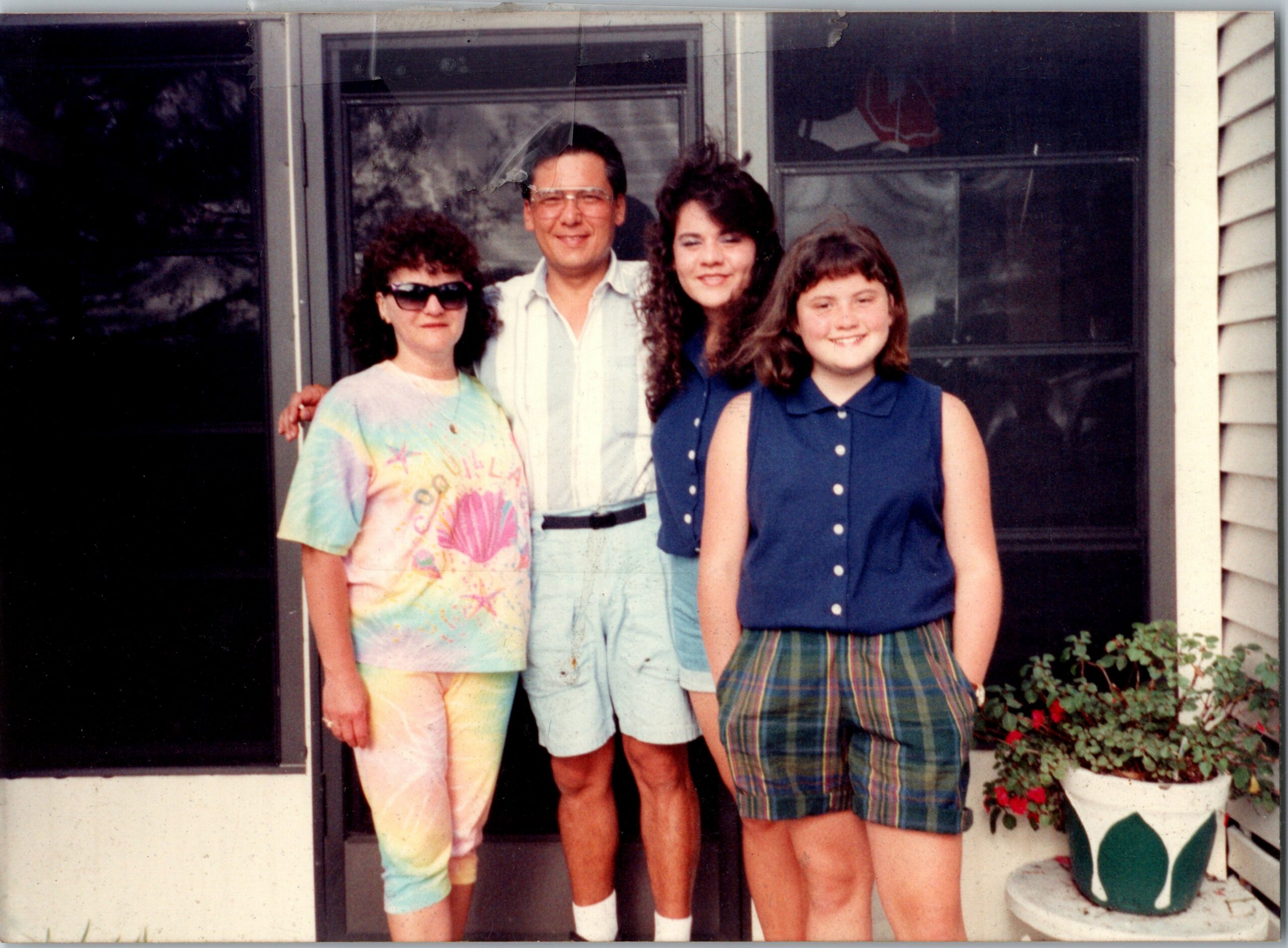 Four people posing on a house front porch: woman in tie-dye shirt and sunglasses, man in light shirt, girl with dark hair in a blue sleeveless top, and girl in a blue plaid skirt; window and flowers visible in background.