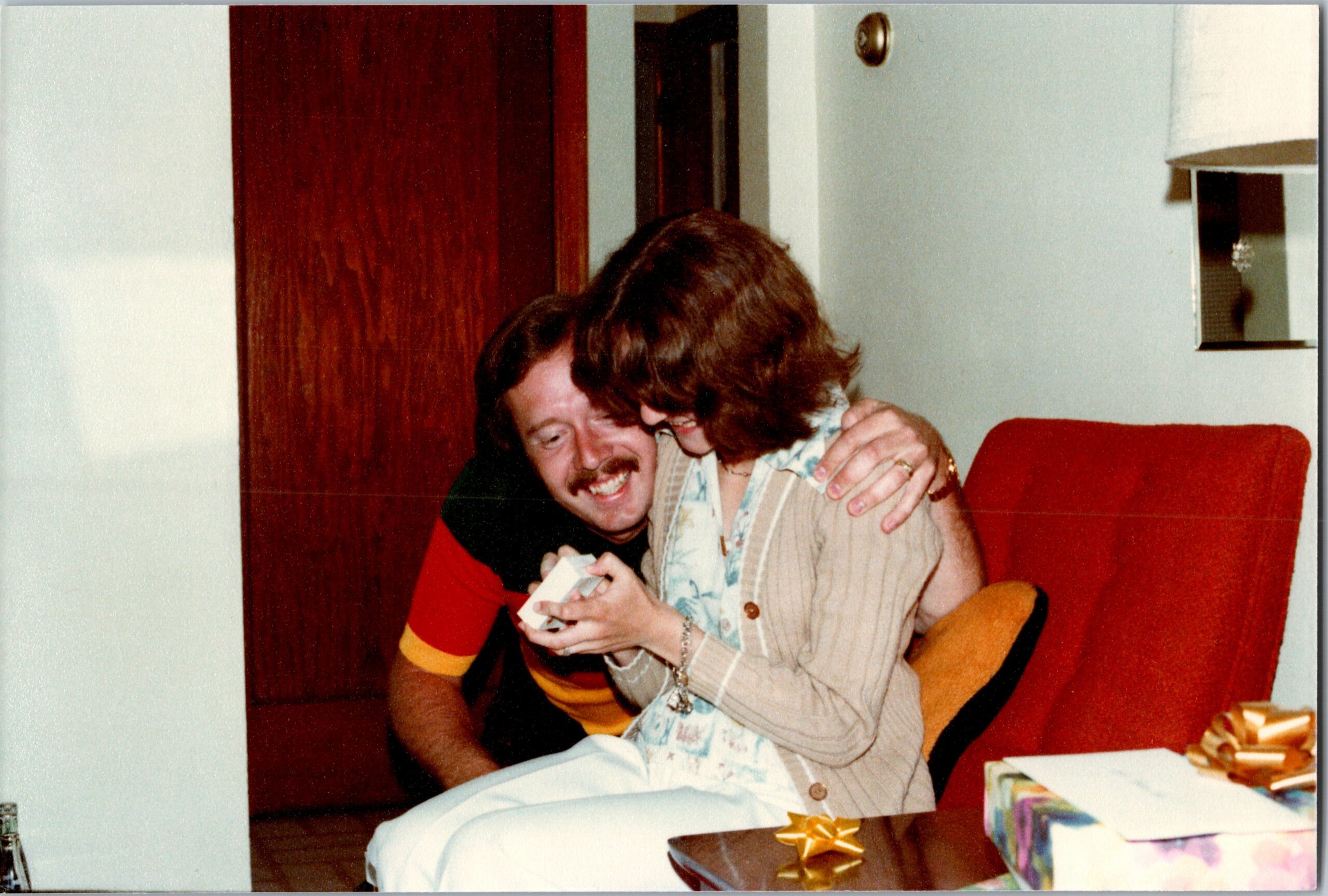 A smiling couple hugs on a red couch as they open a card, with wrapped gifts on a table nearby in a cozy living room.