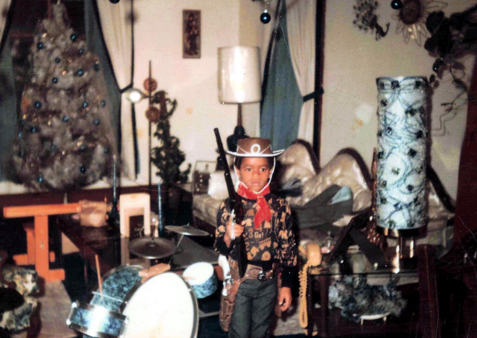 Child in a cowboy hat and red scarf posing with a prop guitar on a small stage, surrounded by holiday decorations and a drum set in the background.