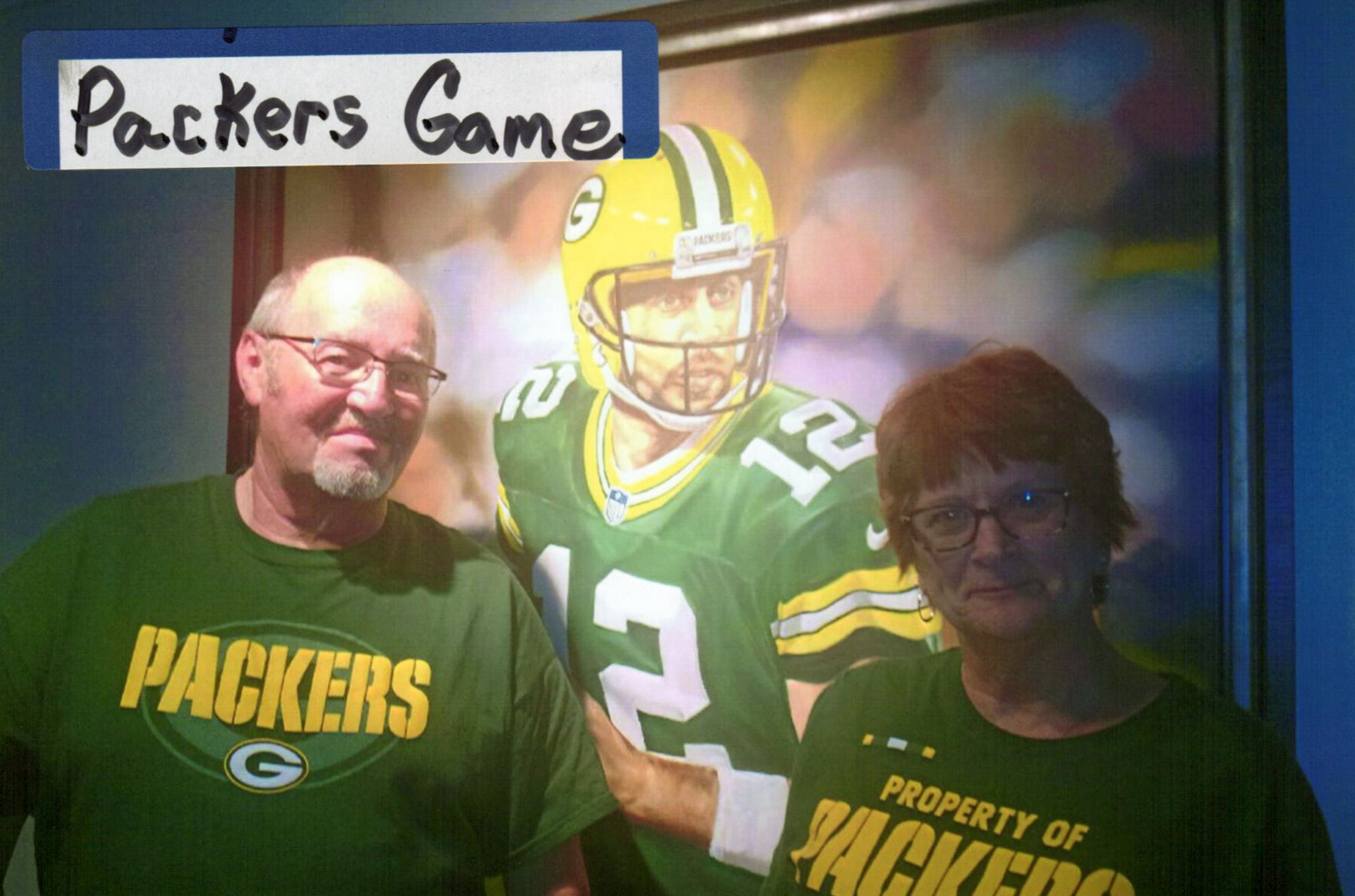Two Packers fans pose beside a life-size image of a Green Bay Packers player, both wearing green Packers shirts.