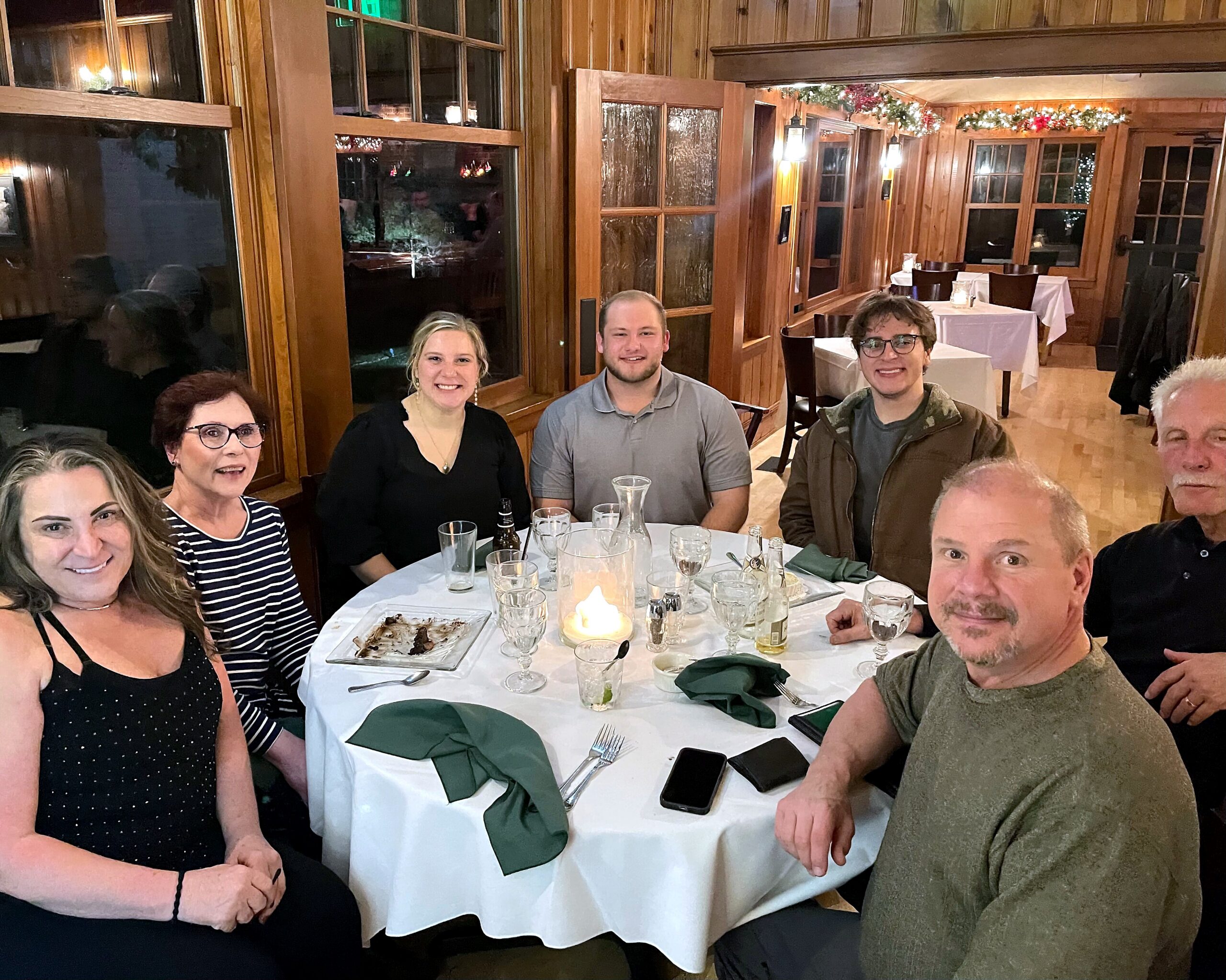Group of friends and family around a candlelit restaurant table, smiling at the camera with plates, drinks, and green napkins on a white tablecloth.