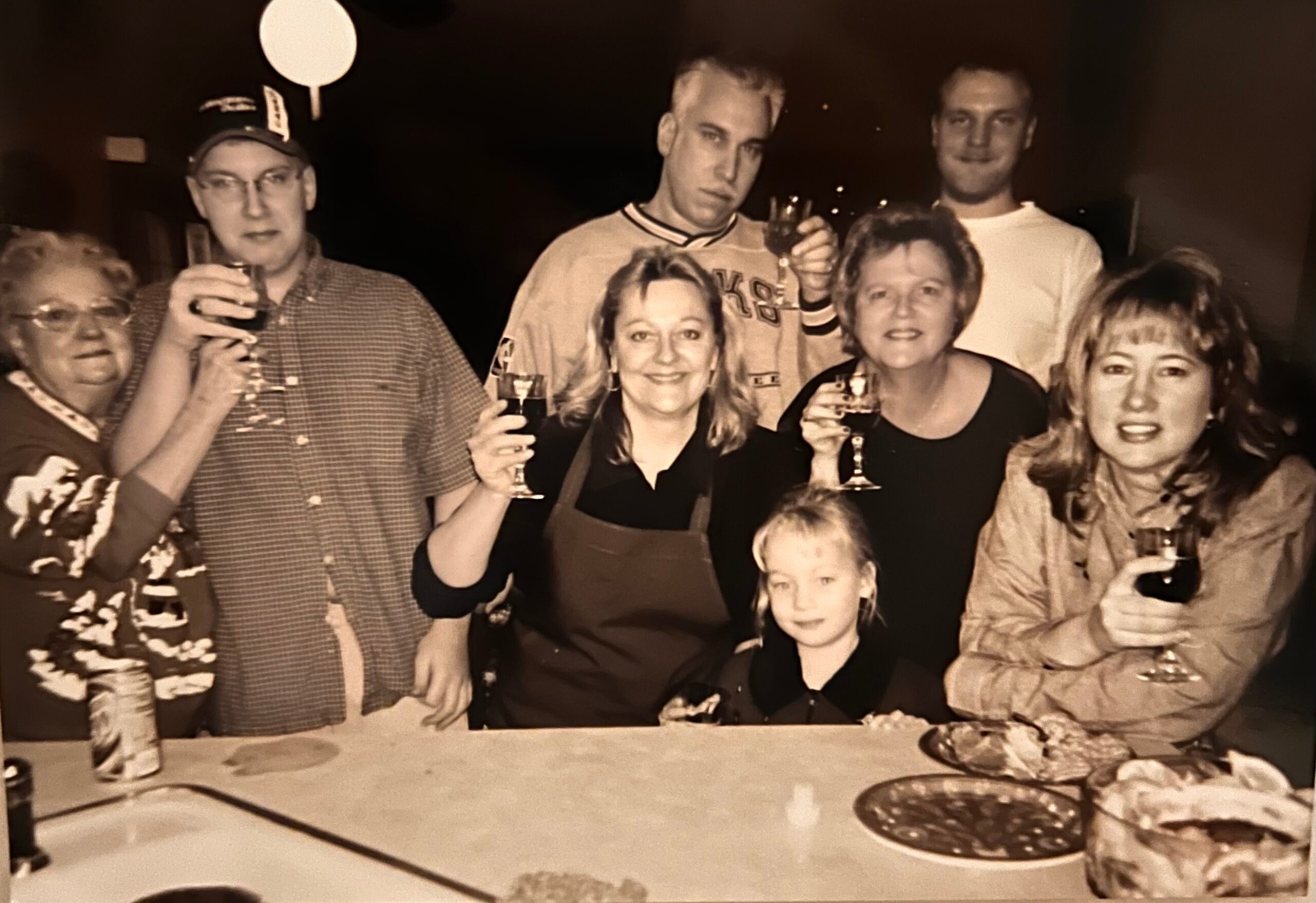 Group of adults and a child posing with drinks at a toast around a table at a party (sepia photo).