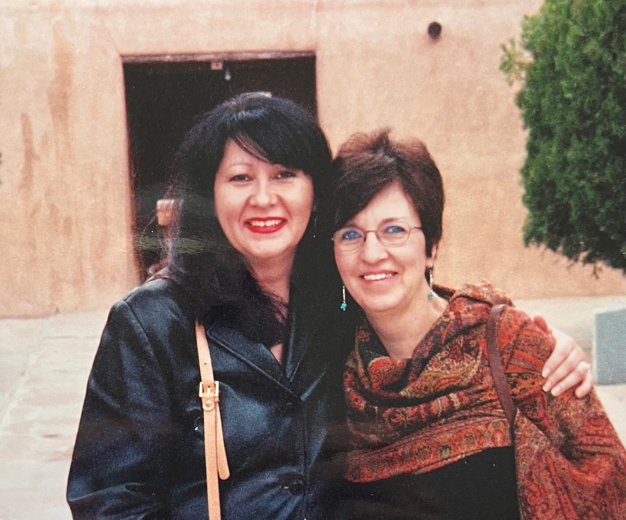 Two women smiling and posing arm-in-arm outdoors, one in a dark coat with a tan purse, the other in a patterned brown dress with turquoise earrings.