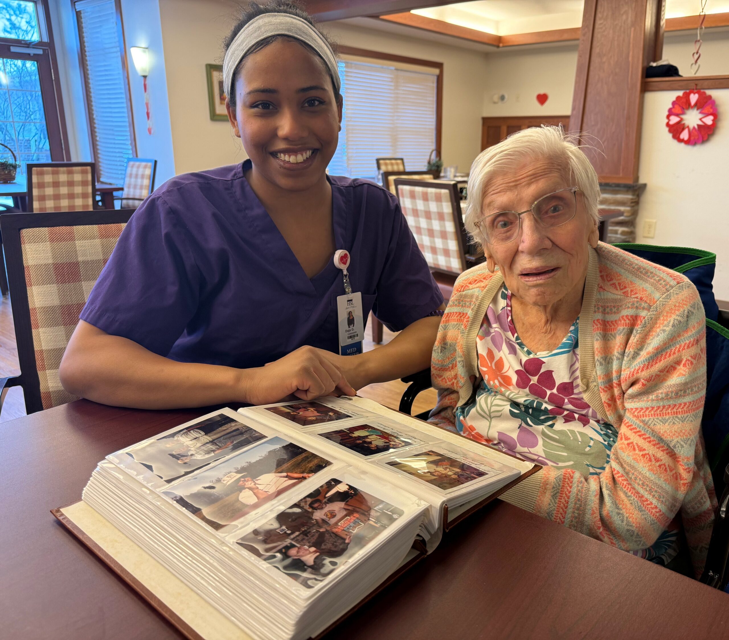 Caregiver in purple scrubs smiles at an elderly woman as they look through photo albums on a table in a dining room setting.