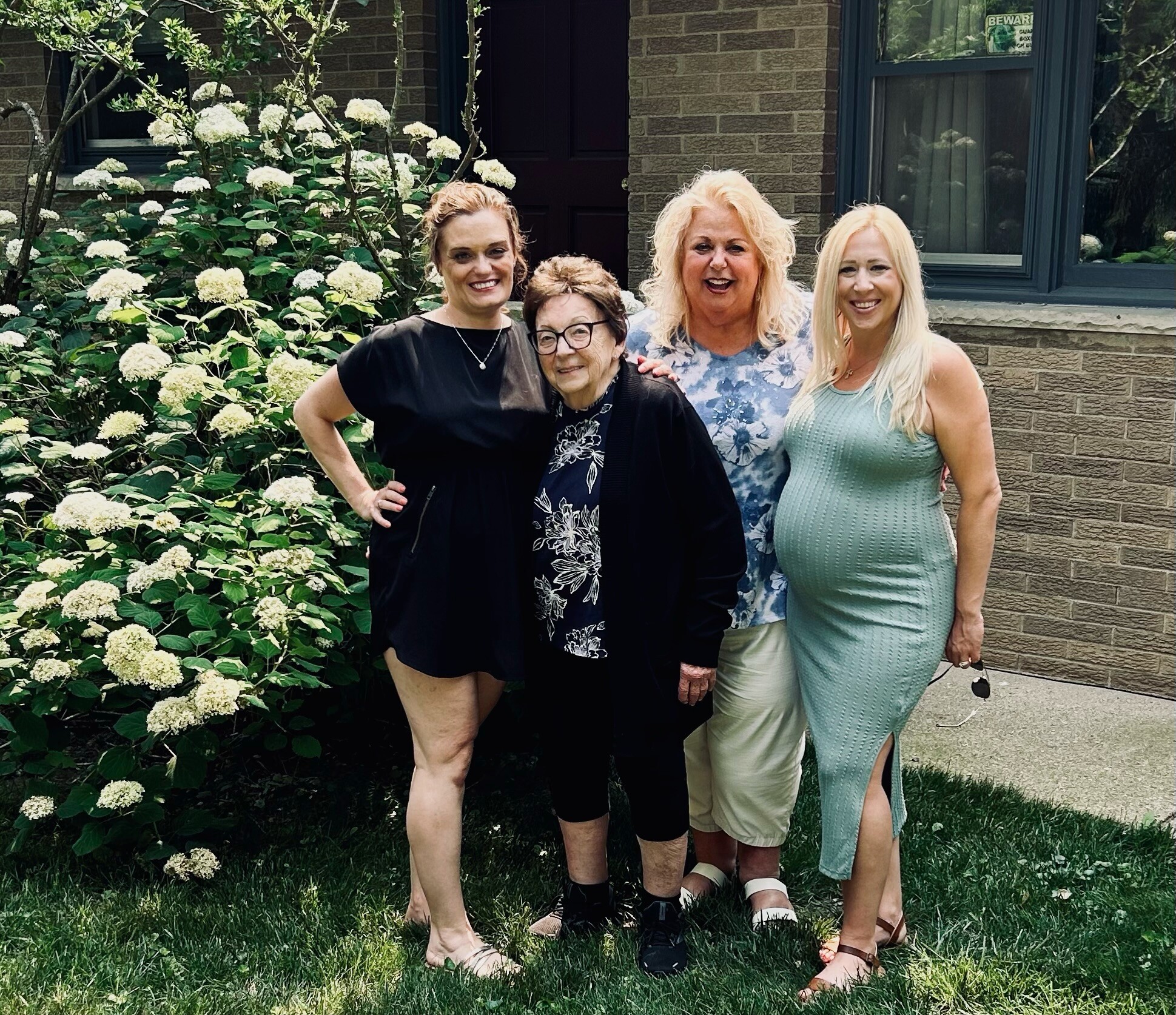 Four women posing and smiling in front of a hydrangea bush by a brick house.