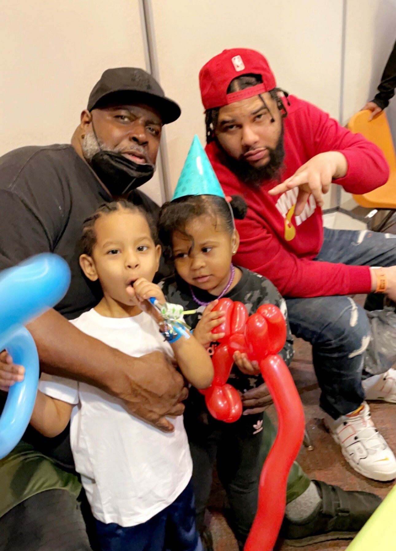 A man in a black cap and mask sits with two children at a party; one child wears a party hat and holds colorful balloon animals while the other holds a red balloon. A second man in a red cap gestures toward the camera in the background.
