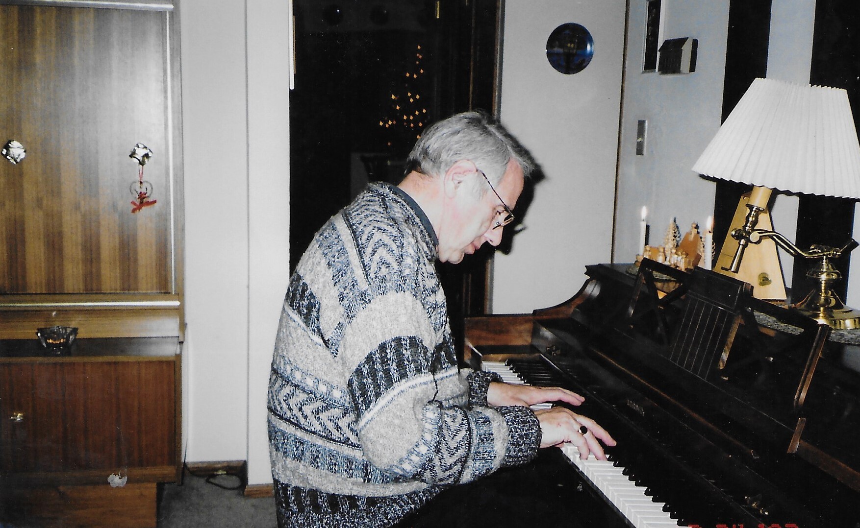 Older man in a patterned sweater playing a piano in a cozy living room, Christmas lights visible outside the window.