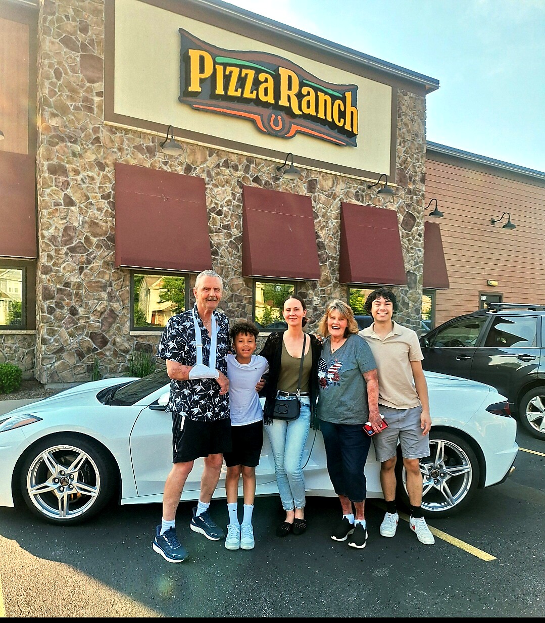 Five people pose in a parking lot in front of a Pizza Ranch restaurant with a white sports car beside them; stone facade and maroon awnings are visible.