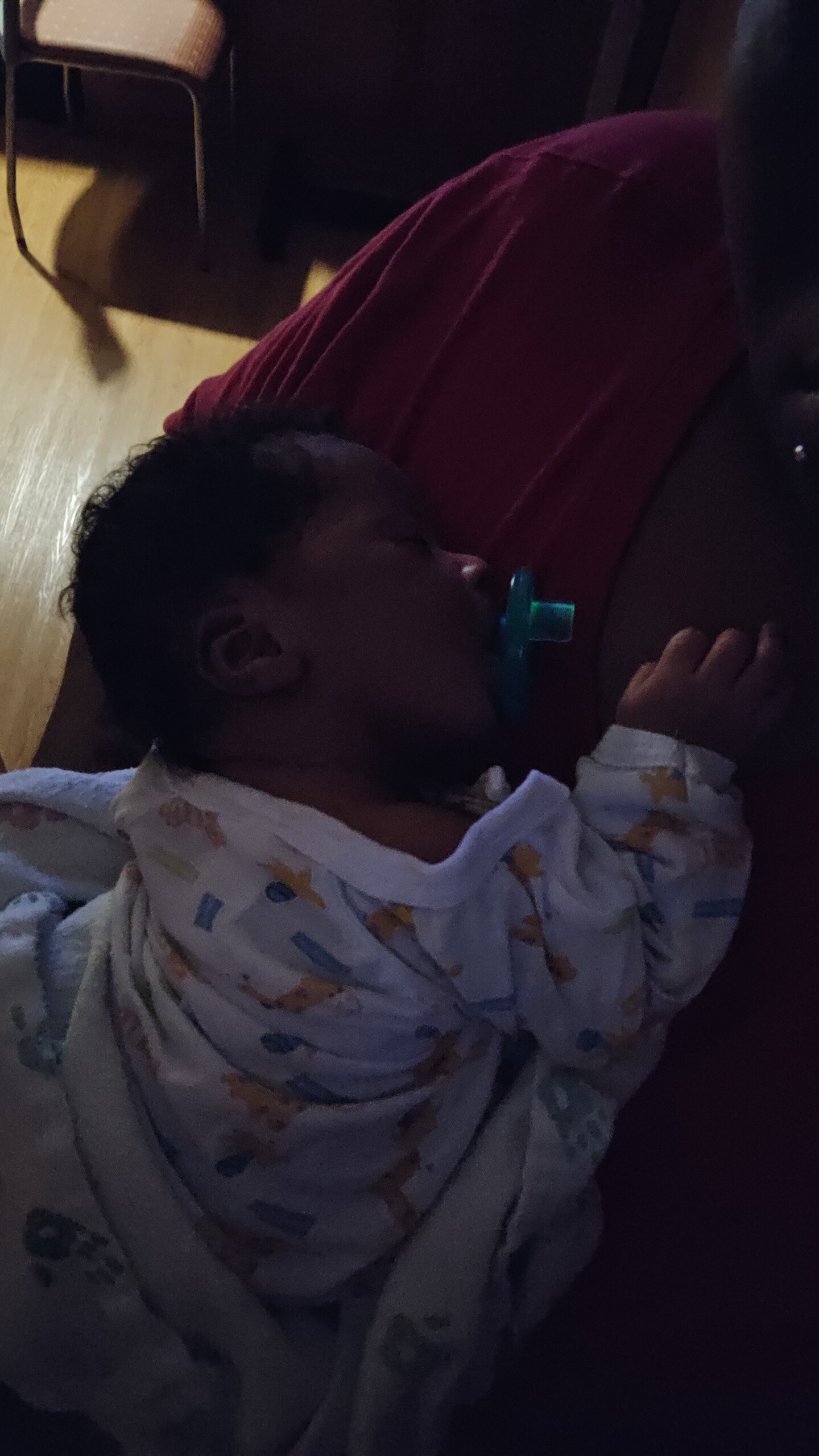 Sleeping baby with a blue pacifier, lying on a red pillow, wearing a pastel hospital gown.