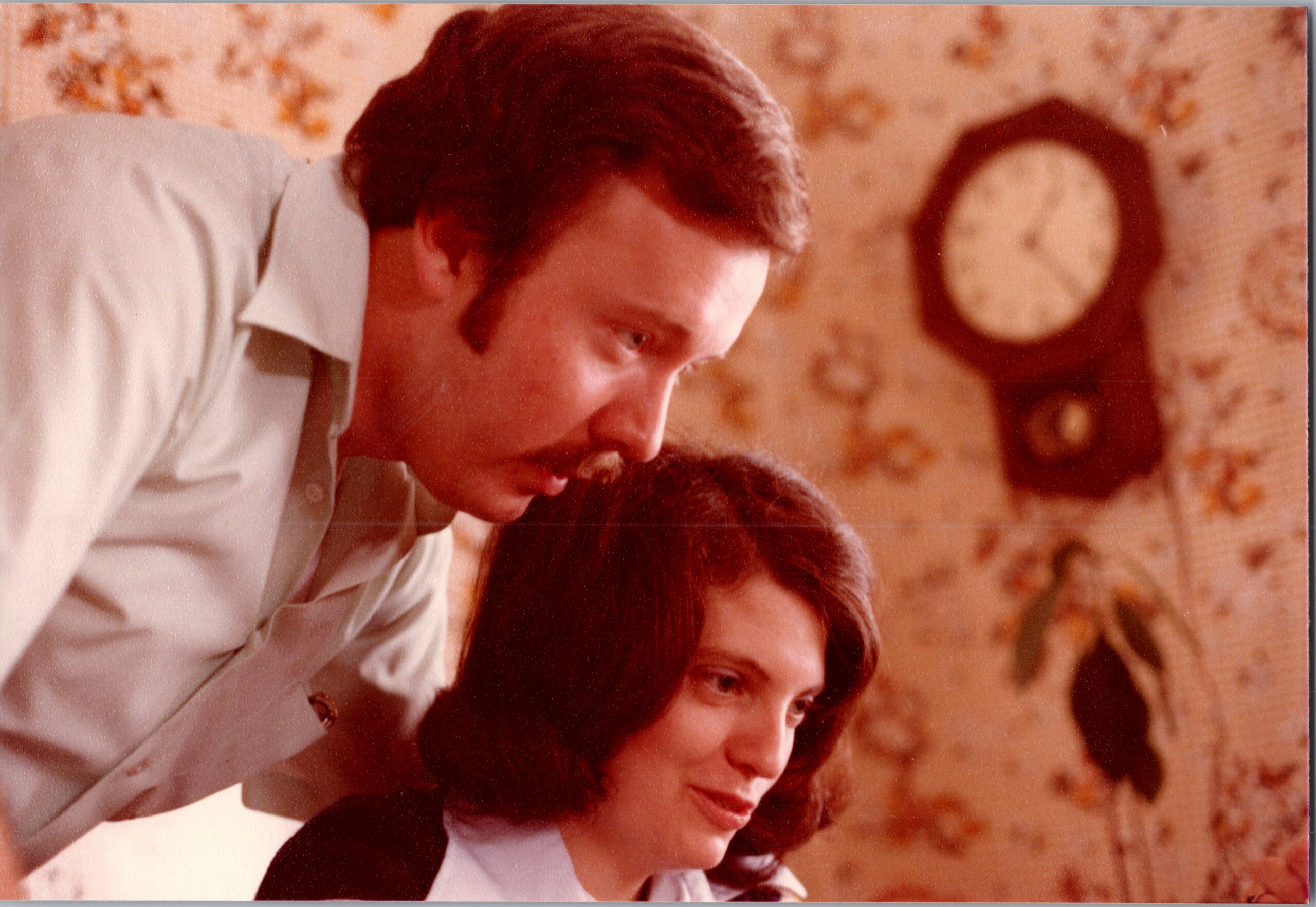 Man leaning over a seated woman with dark hair; floral wallpaper and a round grandfather clock in the background.