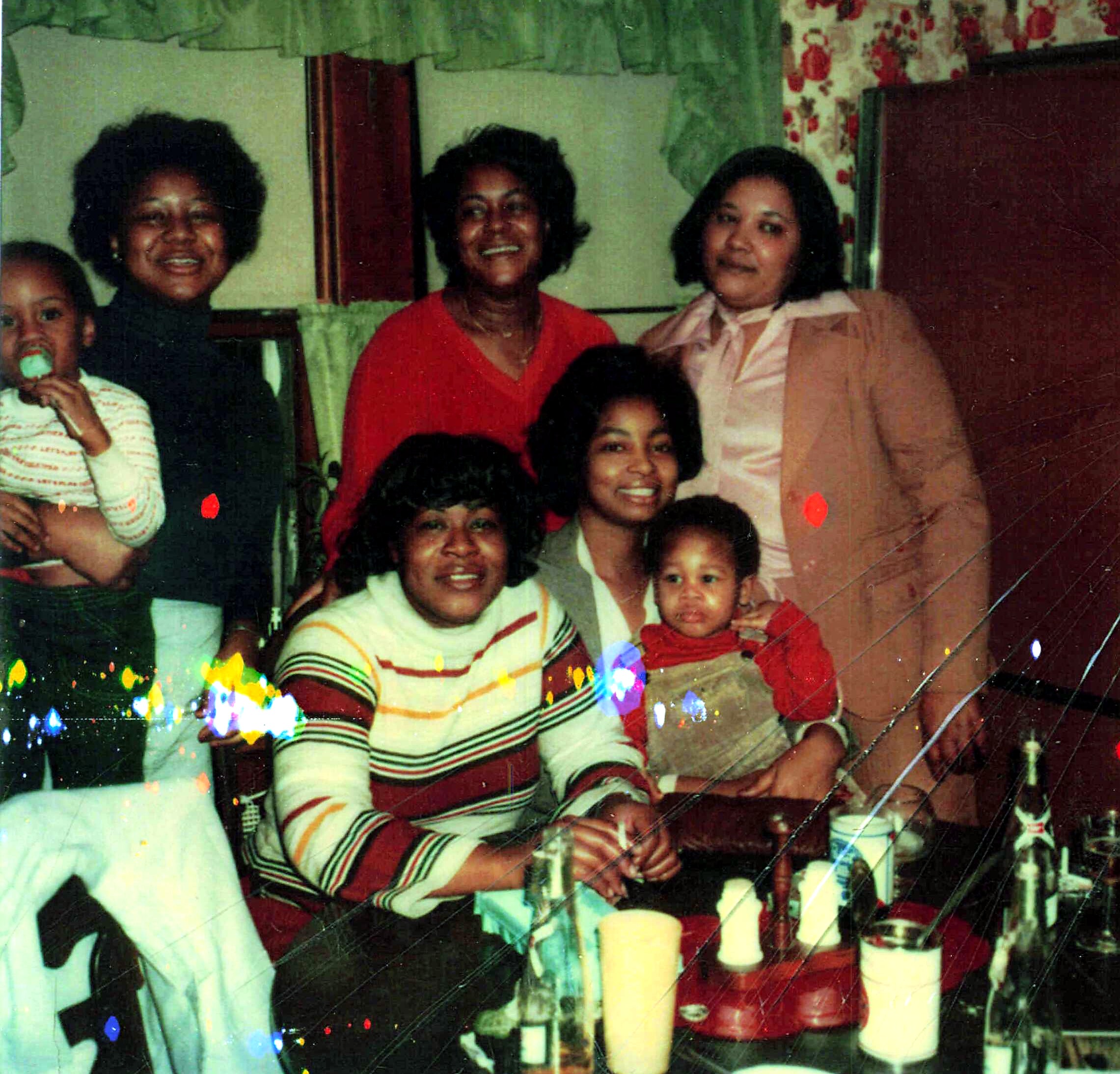 Group of smiling adults and children posing together at a family party around a table with drinks and a cake in a home setting.
