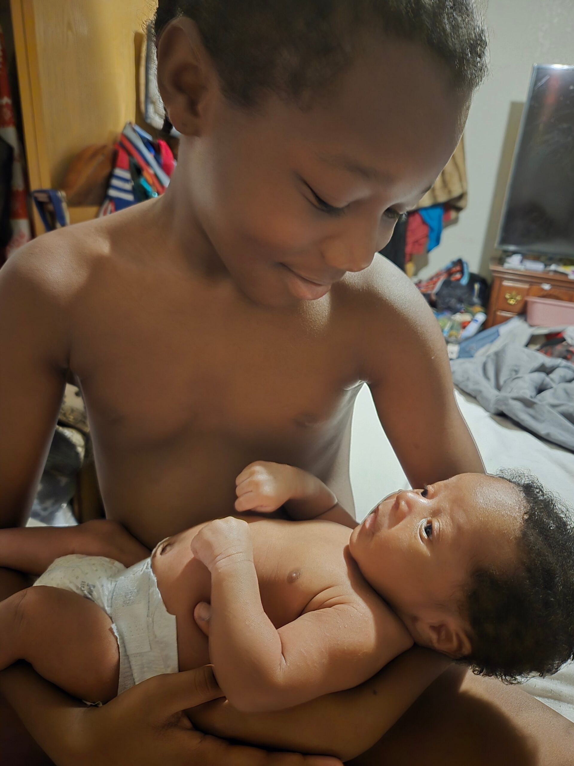 Older boy gently cradles a baby in his arms on a bed, both shirtless in a cluttered room with clothes in the background