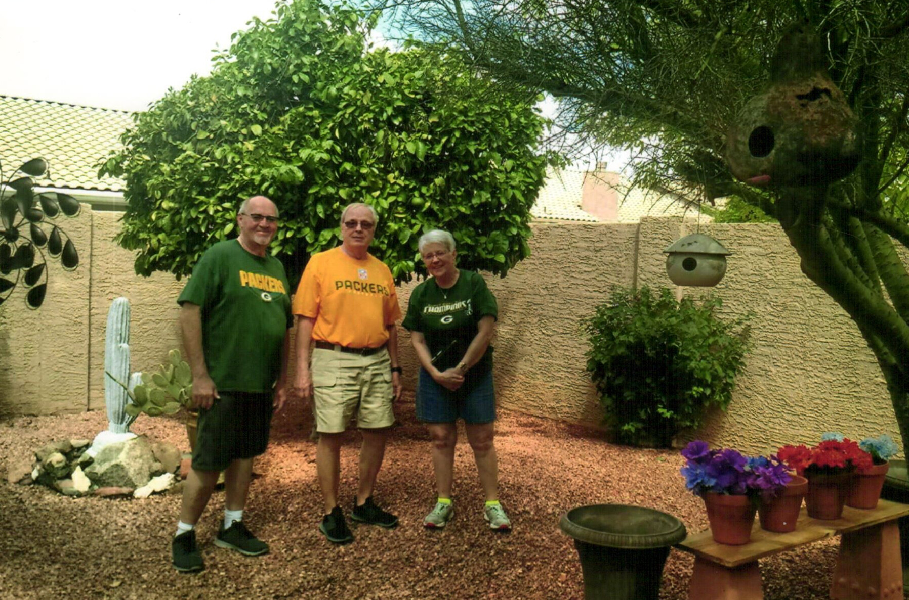 Three adults stand in a desert backyard, wearing Green Bay Packers shirts, with a cactus and greenery in the background.
