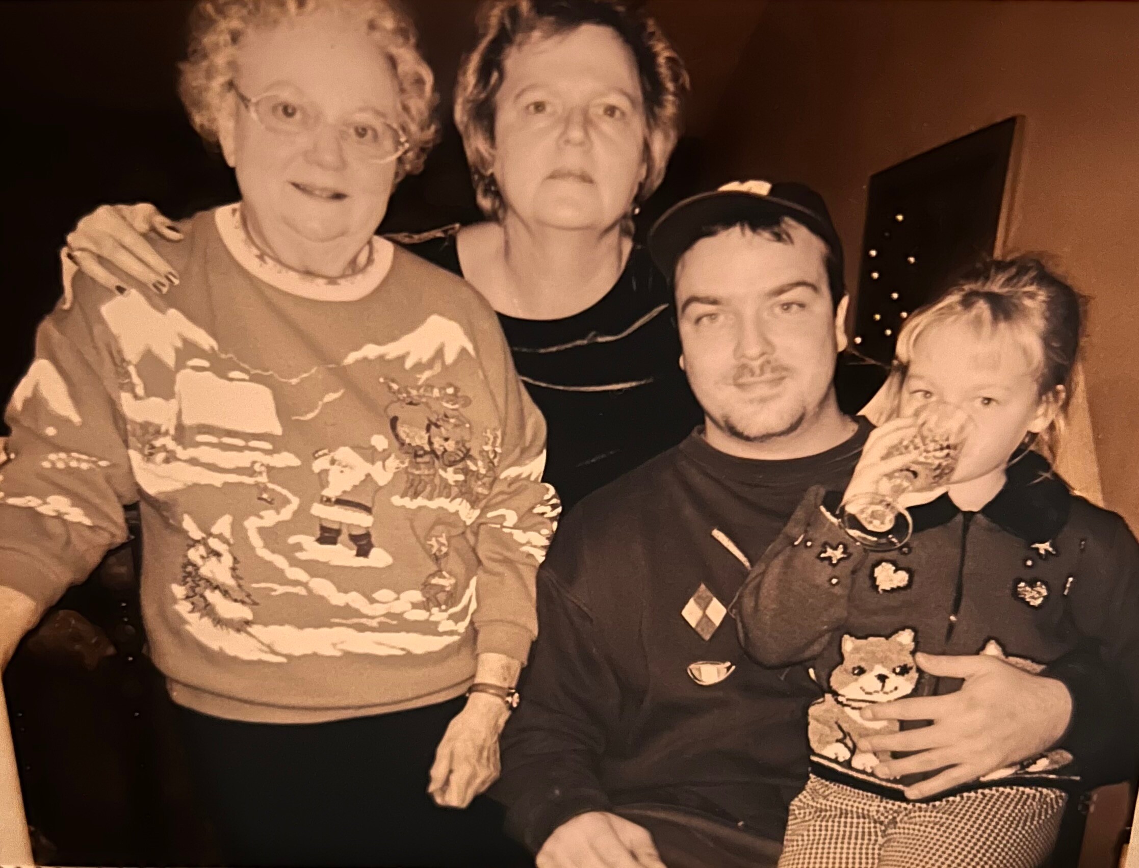 Group portrait of four people: an older woman in a patterned sweater on the left, a woman in the middle, a man with facial hair, and a child on the right holding a small dog.