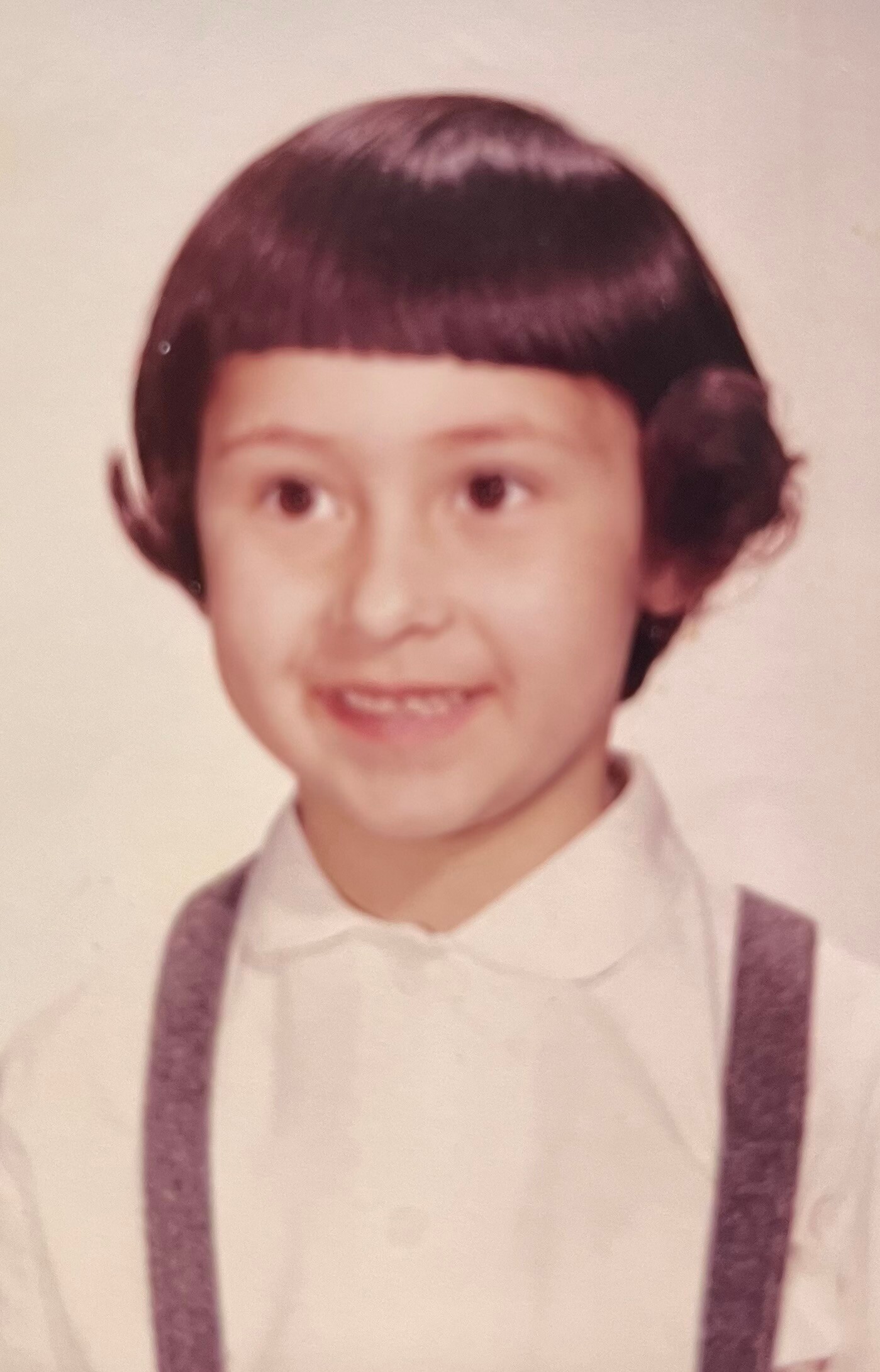 Young child with a dark bowl haircut, white shirt, and purple suspenders, smiling in a vintage studio portrait.