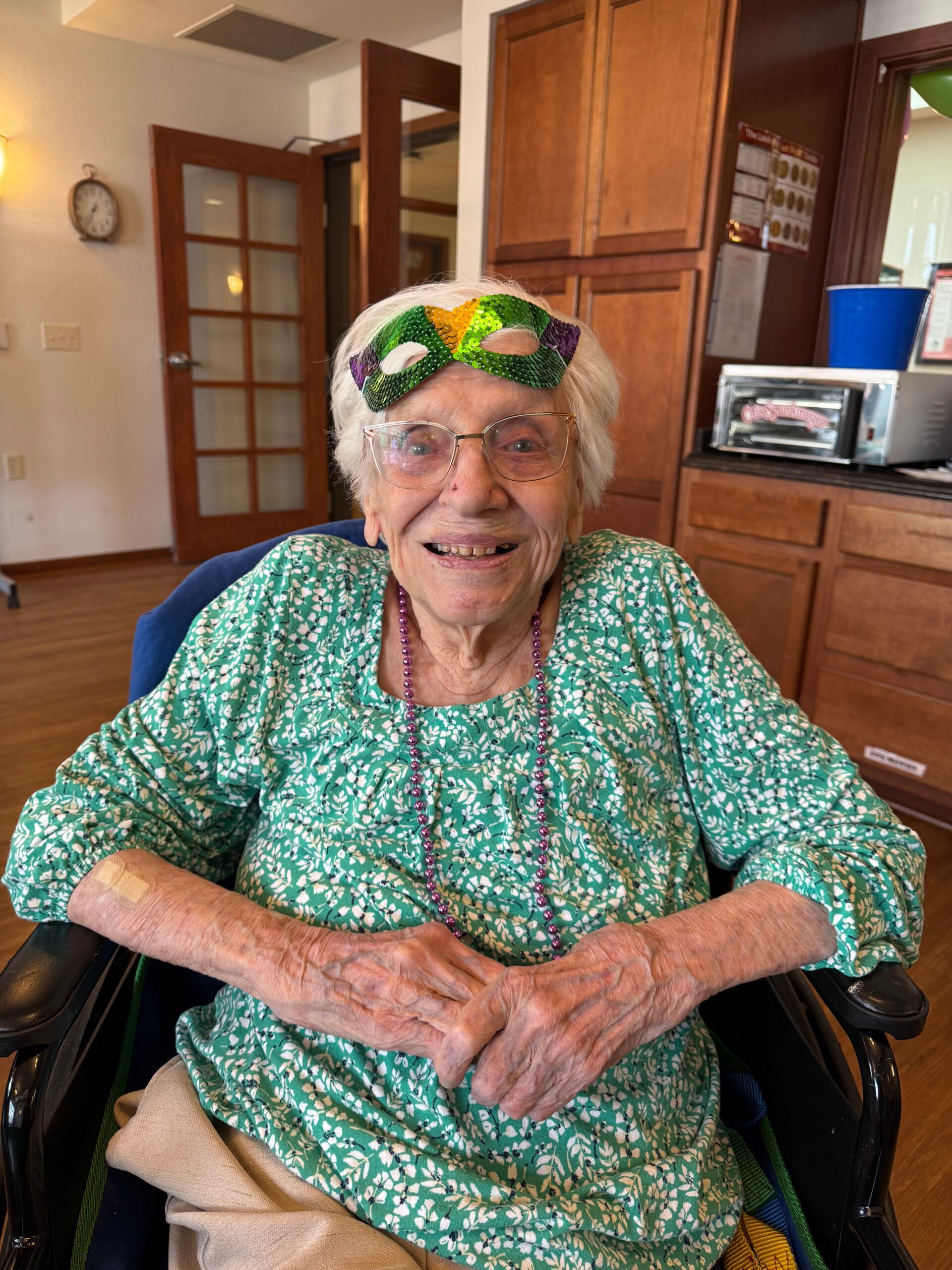 Smiling elderly woman in a wheelchair wearing a green floral blouse with a colorful sequined mask on her forehead.